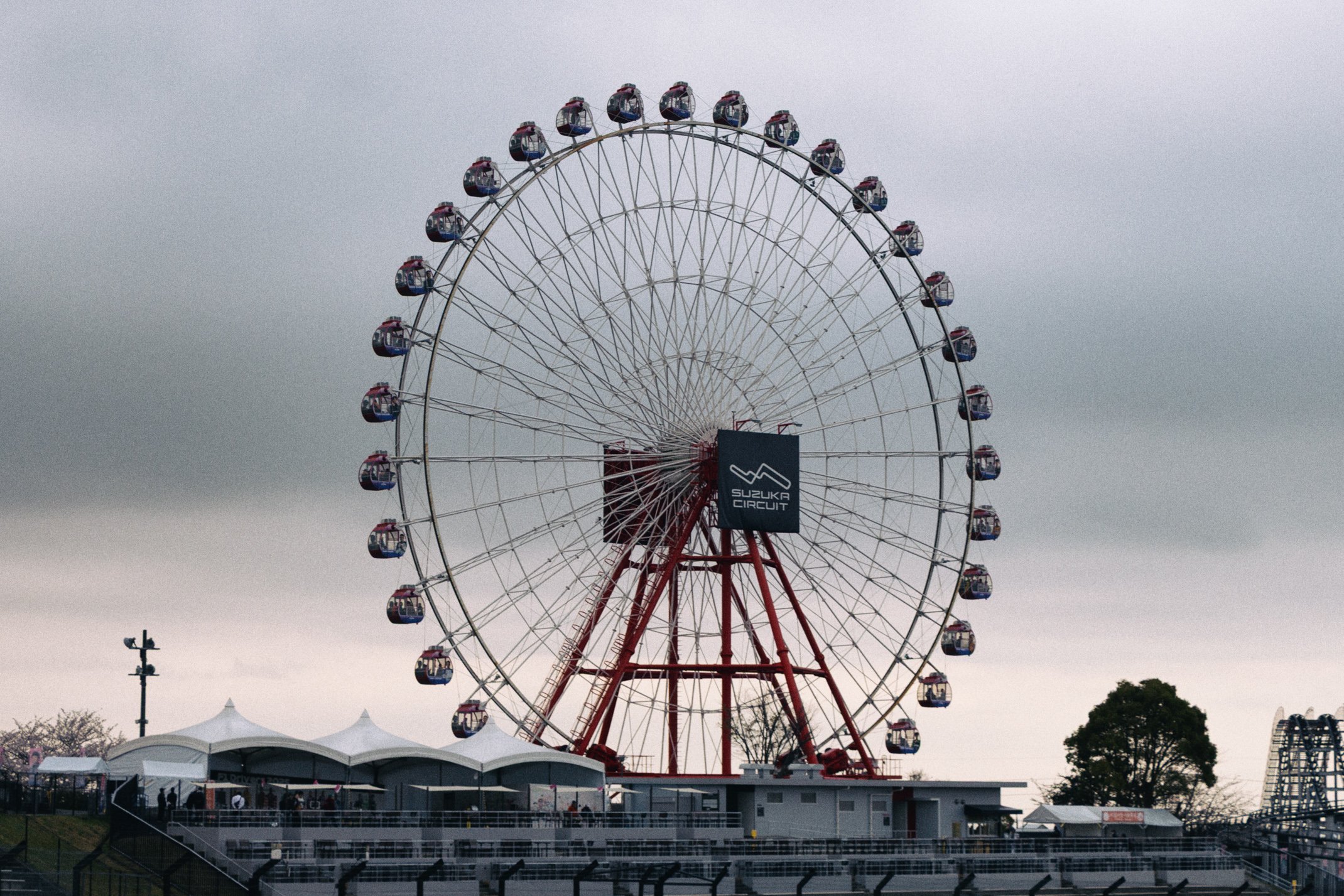 A large Ferris wheel at Suzuka Circuit with multiple cabins, set against a cloudy sky and some tents and trees at the base.
