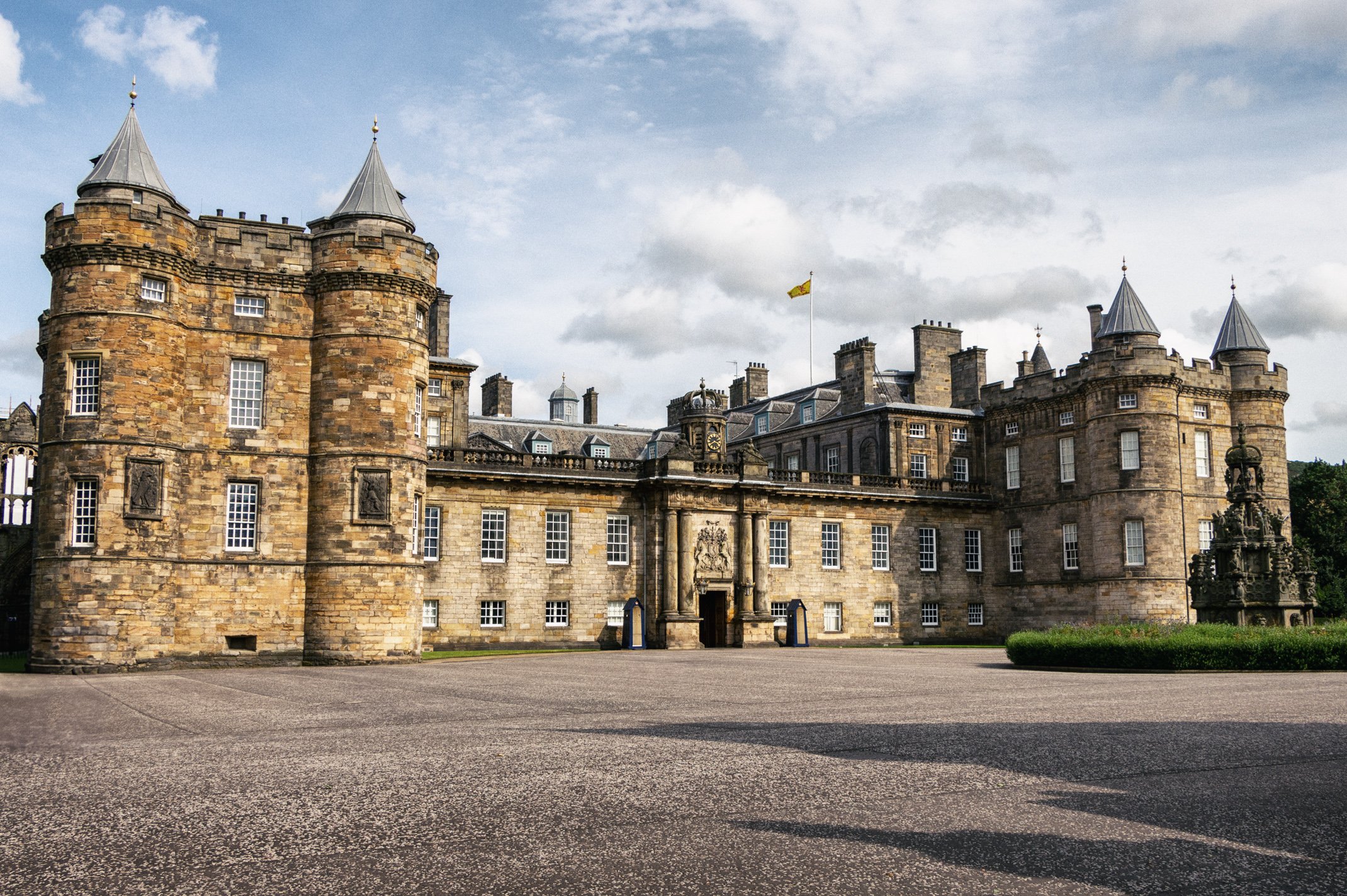 View of a historic stone castle with turrets, windows, and a flag flying on top, set against a partly cloudy sky.
