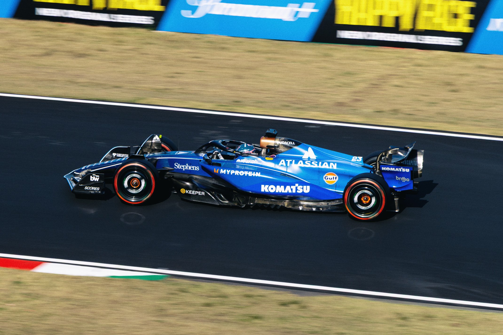 A blue Formula 1 race car on a racetrack, featuring sponsorship logos such as ATLASIAN, KOMATSU, Gulf, and others, with driver in helmet behind the wheel.