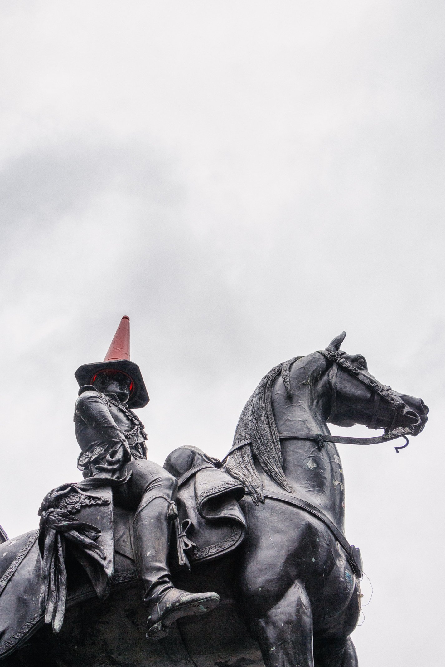 Close-up of a metallic statue of a mounted soldier wearing a traffic cone helmet against a cloudy sky.