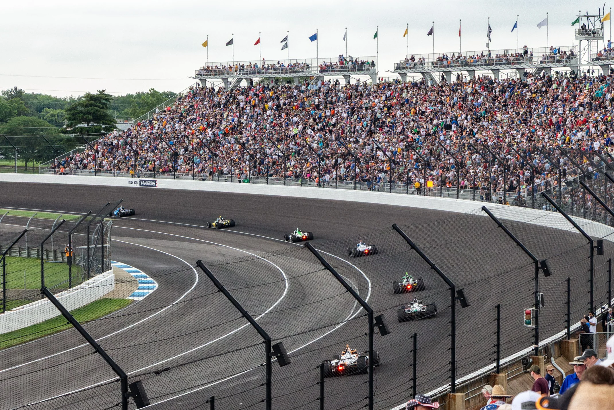 A crowded race track with multiple race cars speeding around the oval, spectators watching from grandstands and fencing.