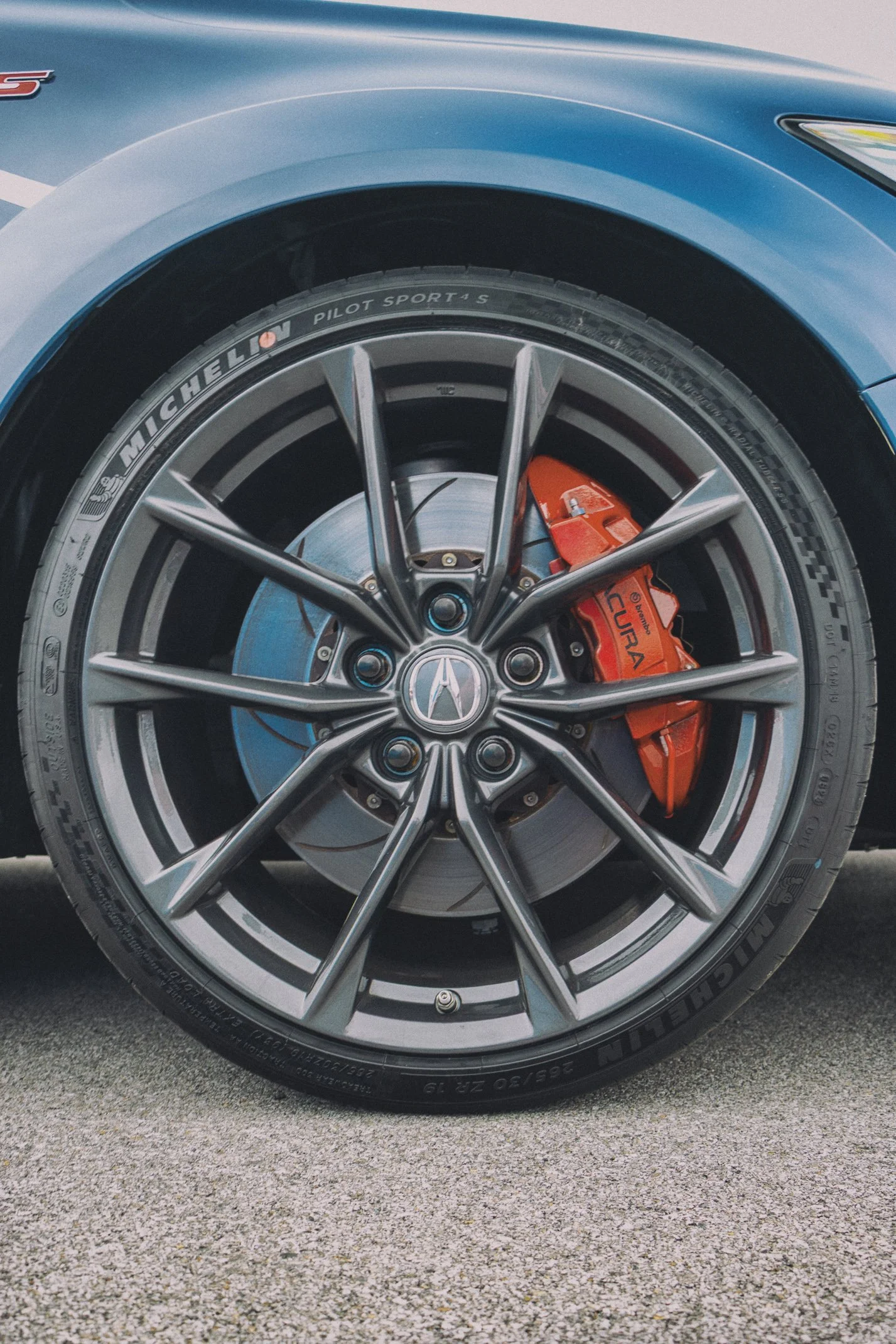 Close-up of a car wheel featuring a Michelin Pilot Sport 4 S tire, a silver alloy rim with a Mercedes-Benz logo, and a visible orange brake caliper with the AMG branding, set on a textured asphalt surface.