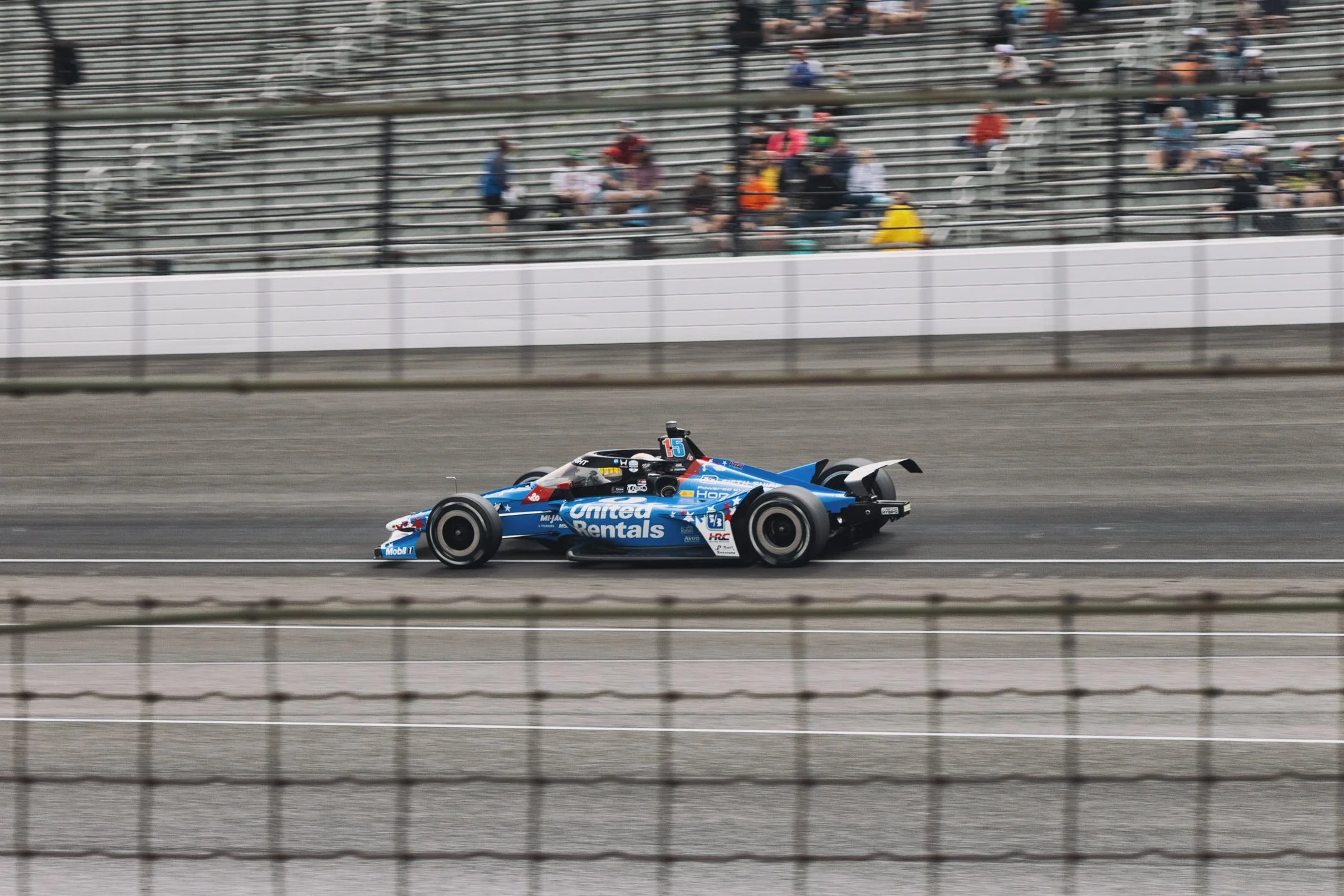 A blue race car on a race track, with spectators behind a fence in the background.