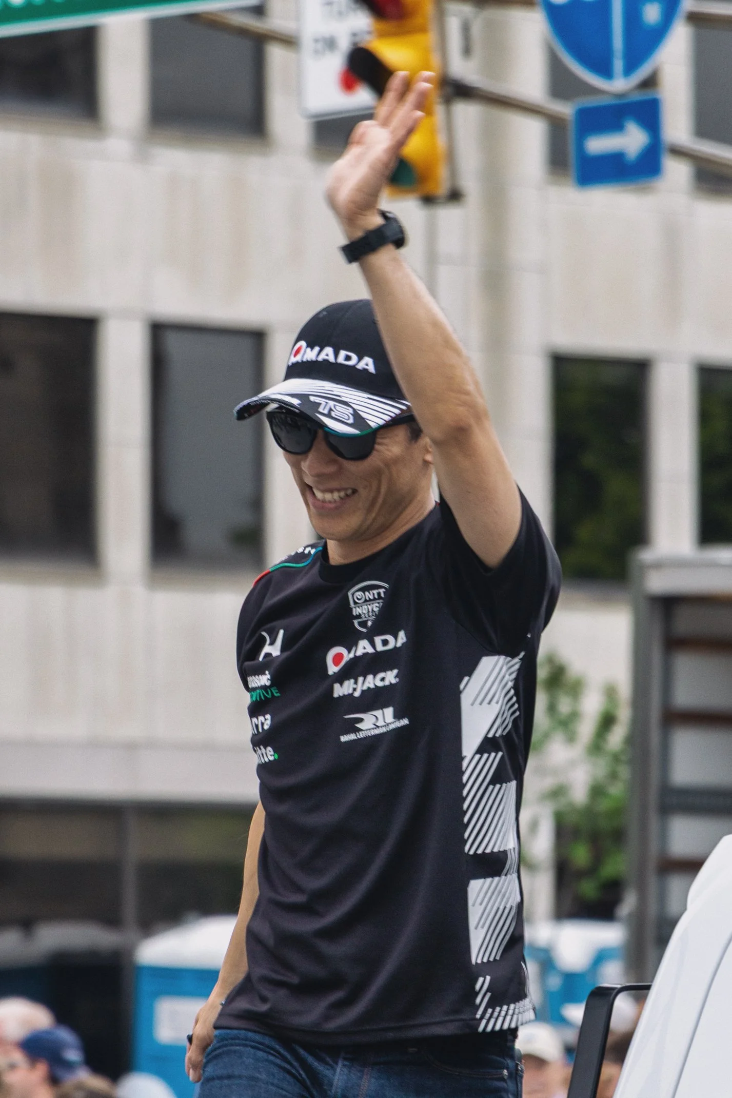 A man wearing sunglasses, a black cap with the logo 'OAMADA' and a black sports jersey with various sponsor logos on it, is raising his hand. The background shows a city street with a building, street signage, and other people.