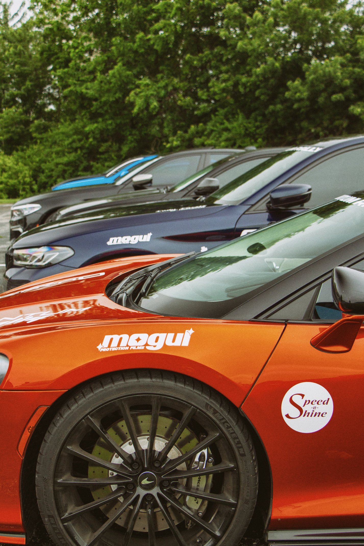 Close-up of a red sports car with black wheels and a white circular sticker that reads 'Speed Shine' on the front fender, parked alongside other cars with a background of green trees.
