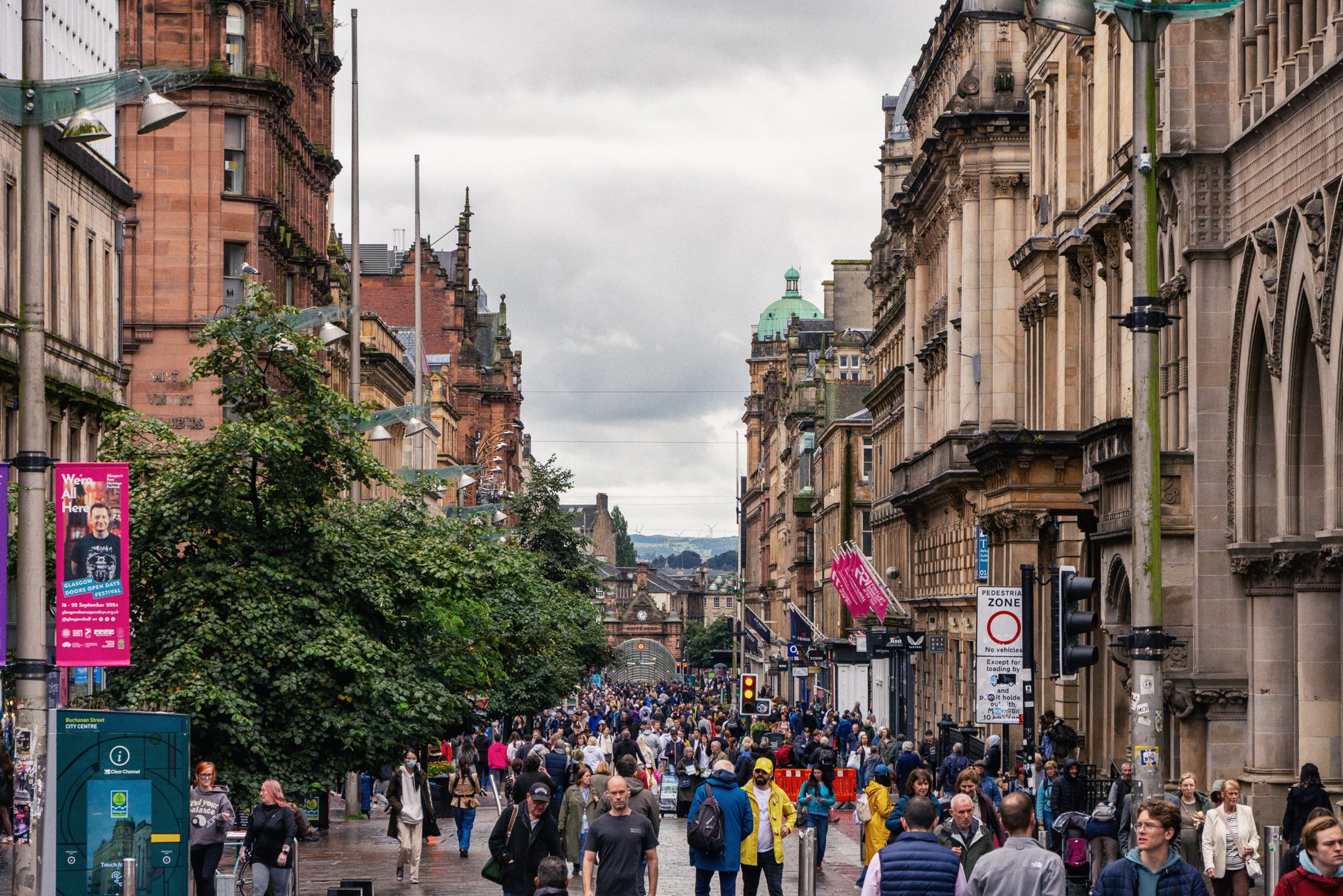Crowded city street with historic buildings, trees, and pedestrians. Overcast sky with a mix of people walking and standing, some in masks, in front of shops and a bus stop.