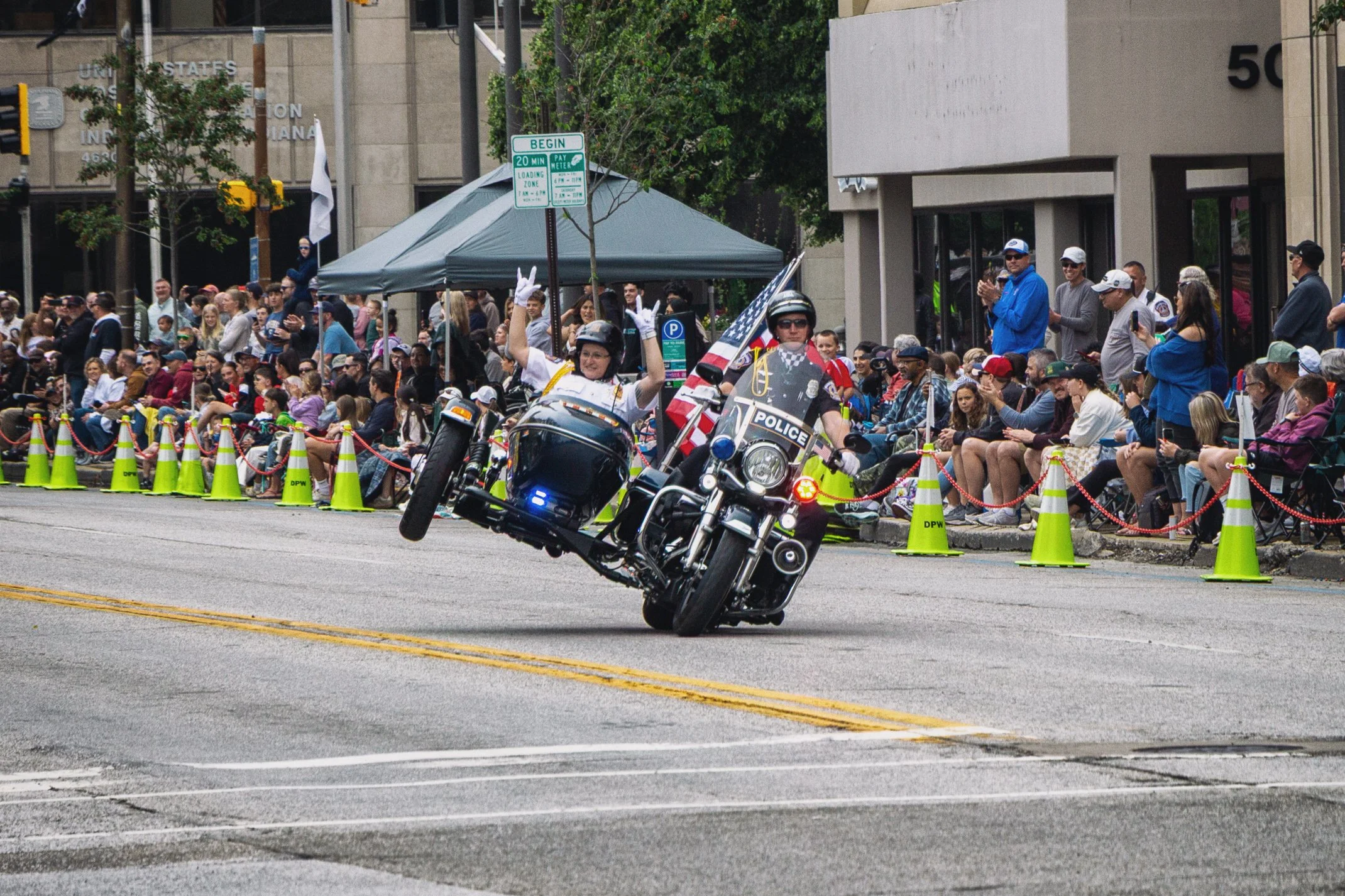 A police motorcycle performing a stunt during a parade with a large crowd of spectators sitting and standing along the sidewalk, some taking photos, and a green tent set up in the background.