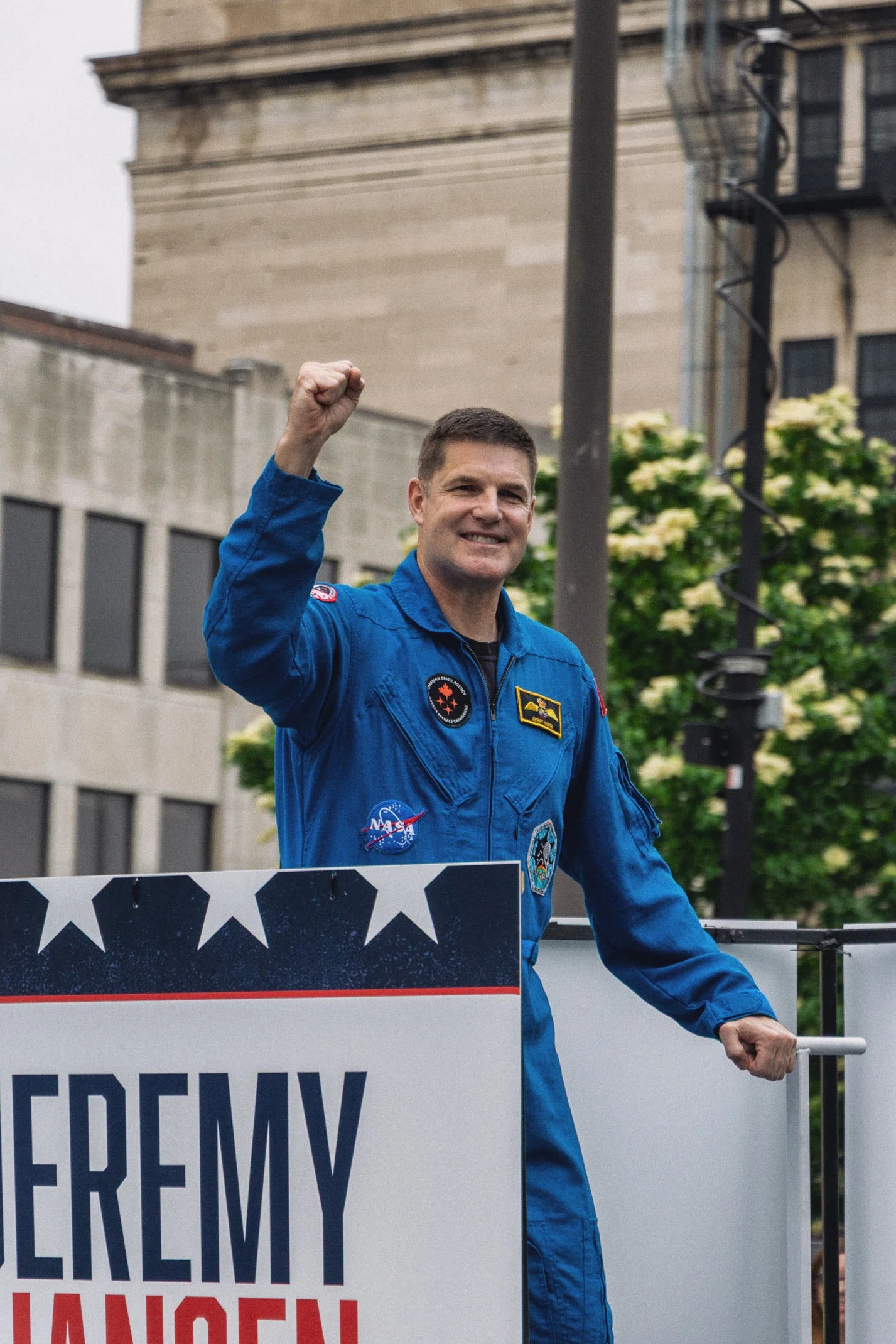 A man dressed in a blue NASA jumpsuit raising his fist at a parade or demonstration, standing behind a sign that partially reads 'U.S. Army'. There are buildings and trees in the background.