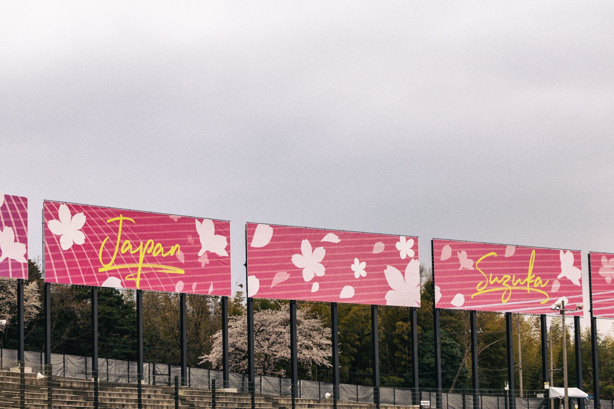 Large pink banners with cherry blossom designs and yellow handwritten-style text displaying 'Japan' and 'Suzuka' on a fence near trees with cherry blossoms, under a cloudy sky.