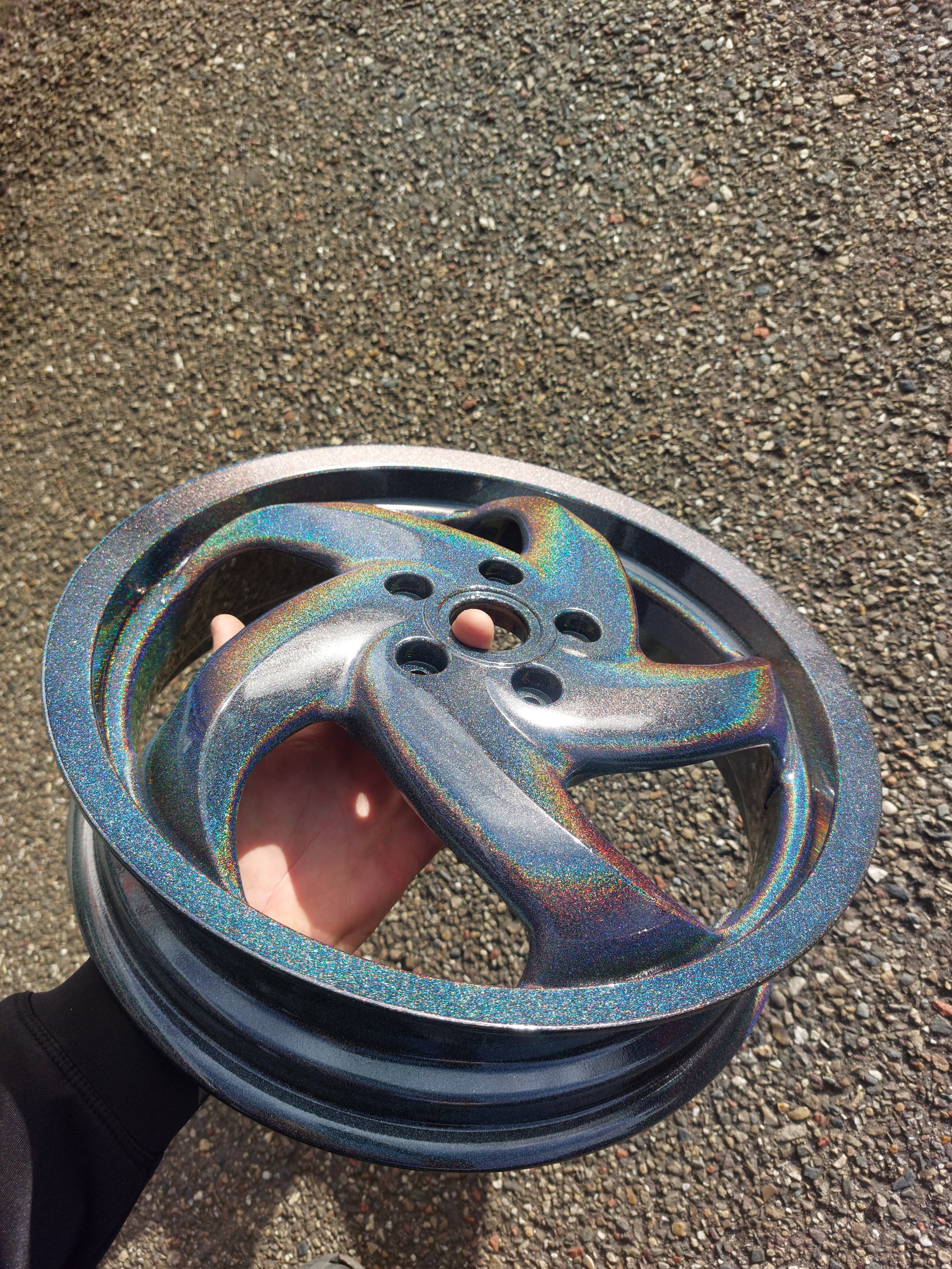 Metalen velg met regenboogachtige glans, vastgehouden door een hand boven een grindoppervlak