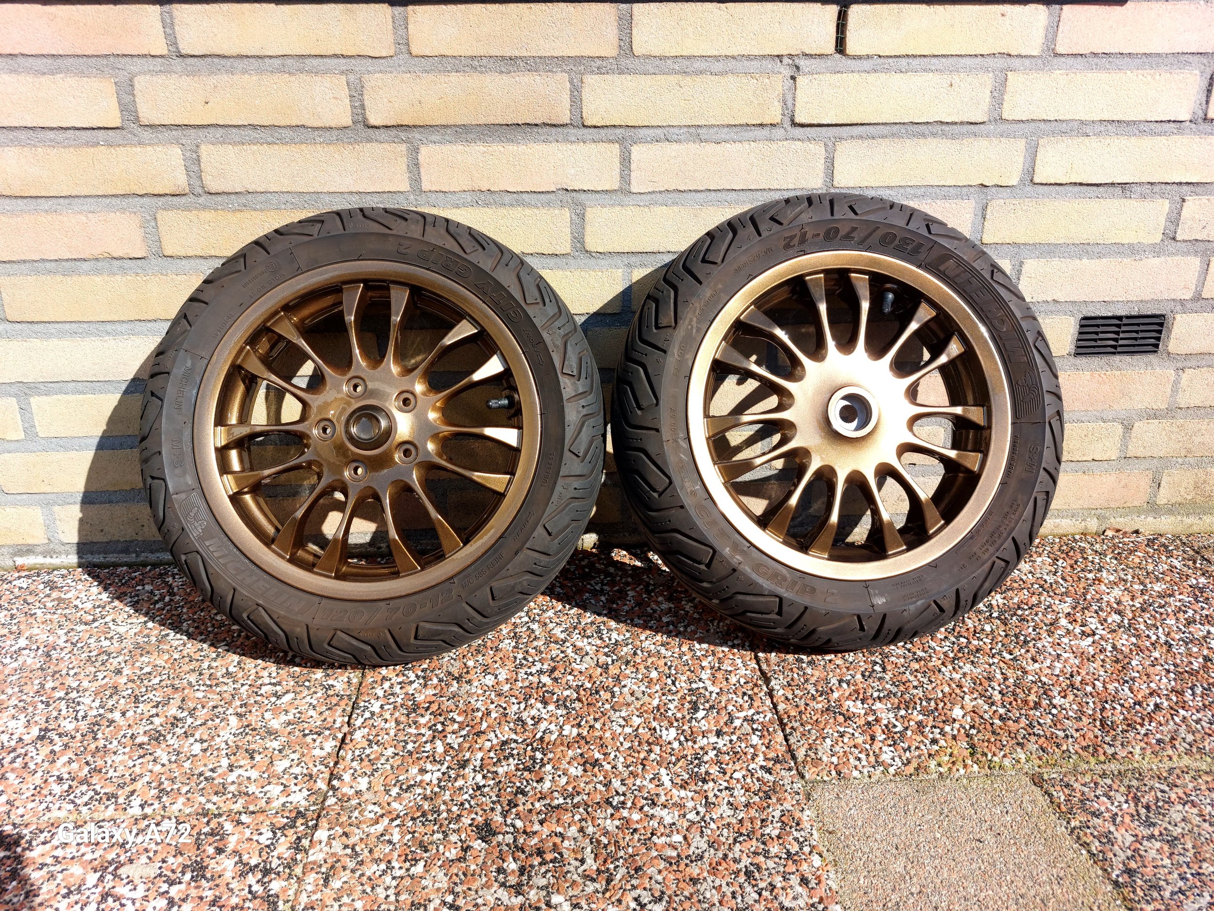 Twee bronzen autovelgen met banden tegen een stenen muur.