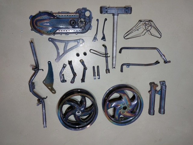 Verschillende onderdelen van een scooter of fiets, waaronder wielen, assen, een frame, en andere metalen componenten, verspreid op een vlakke ondergrond.