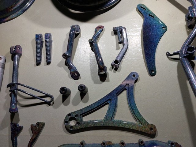Verschillende metalen componenten met een iriserende afwerking, gerangschikt op een tafel. De onderdelen variëren in vorm en grootte en lijken afkomstig te zijn van een mechanisch apparaat of voertuig.