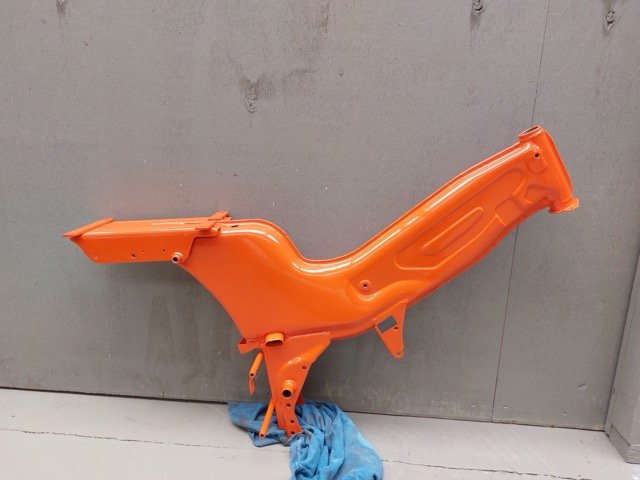 Oranje metalen frame van een bromfiets tegen een betonnen achtergrond. Er ligt een blauwe doek op de grond.