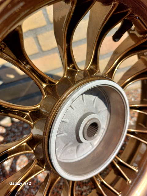 Close-up van een goudkleurig wiel met complexe spaken, waarschijnlijk onderdeel van een voertuig zoals een motorfiets, met een bakstenen muur op de achtergrond.