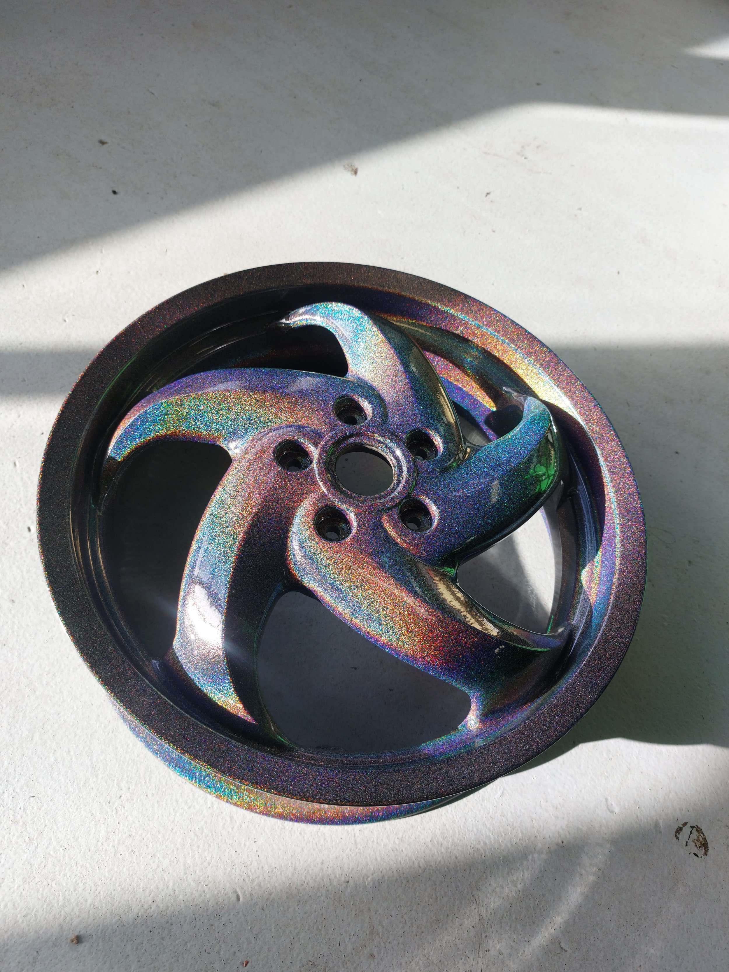 Metaalachtige velg met regenboogafwerking op een lichte ondergrond.