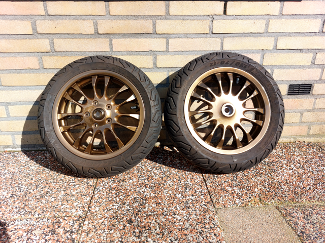 Twee bronzen autovelgen met banden tegen een bakstenen muur op een grindvloer.