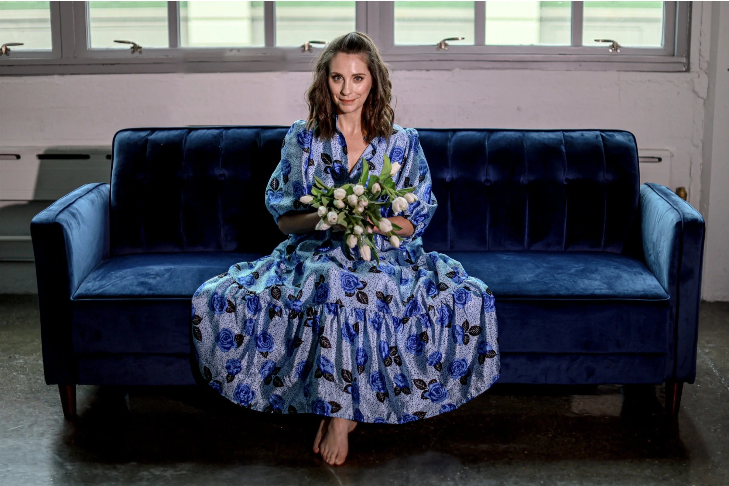 A woman sitting on a navy blue velvet couch, holding a bouquet of white flowers, in a room with large windows and white walls.