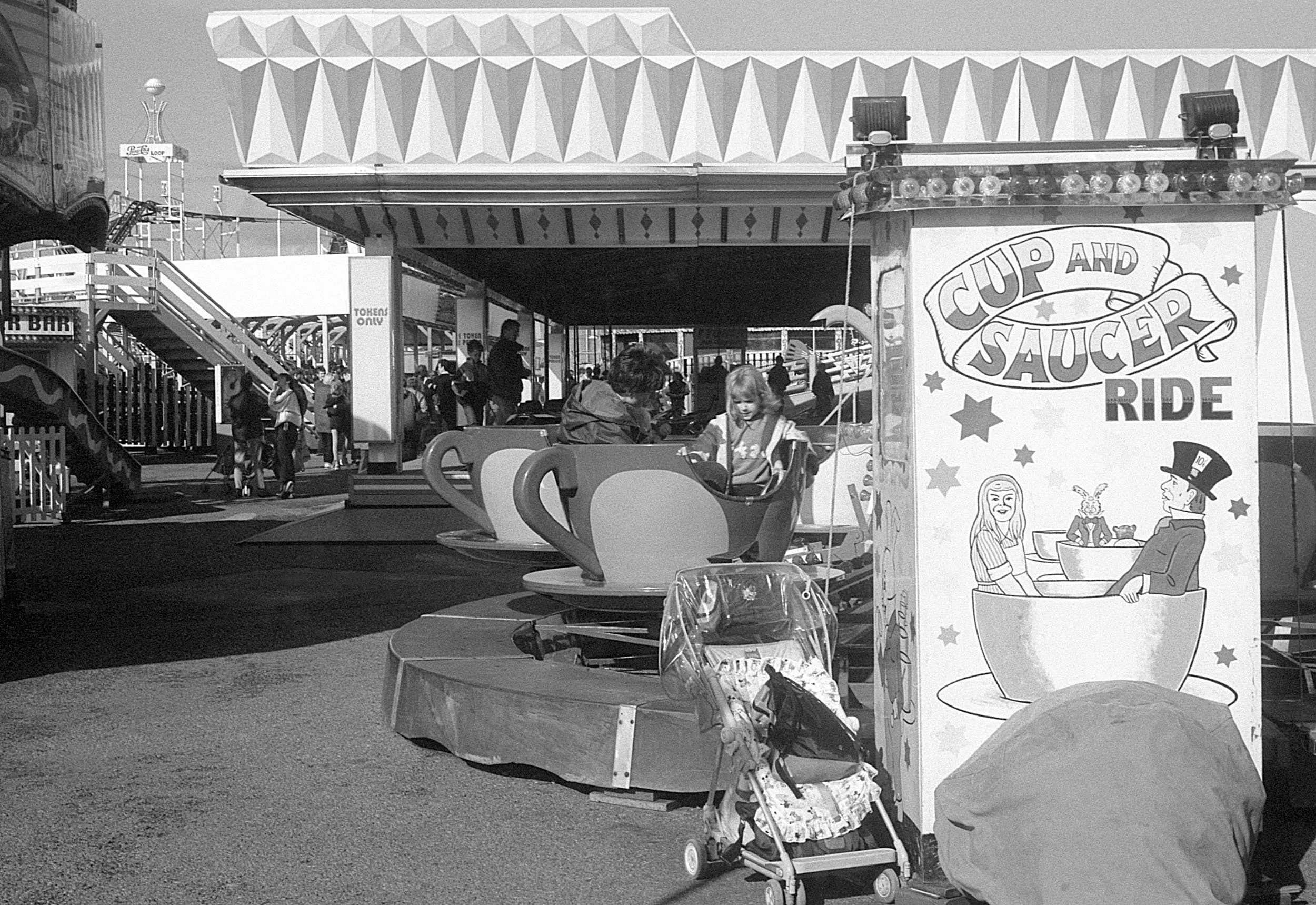 A scene at an amusement park with children playing and riding amusement rides, including a tea cup ride called 'Cup and Saucer Ride,' with a stroller and various rides and attractions in the background.