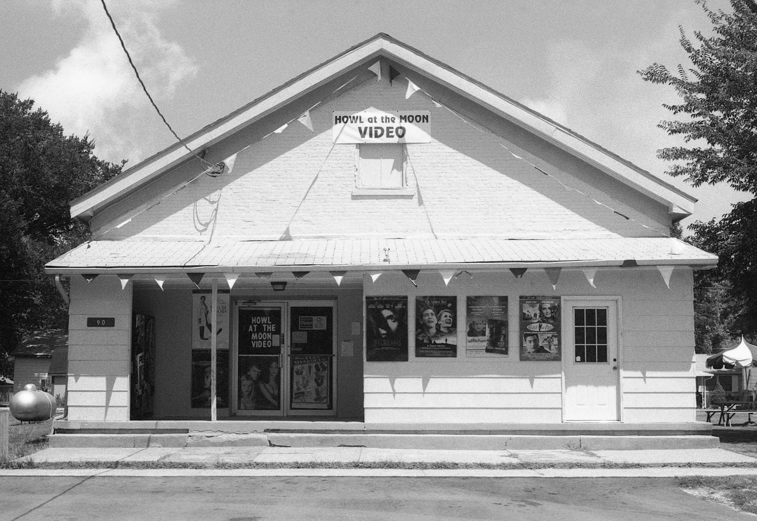 A small, white wooden building with a gabled roof, signs on the front and above the entrance, and posters on the wall, indicating it is a video rental store called 'Howl at the Moon Video'.