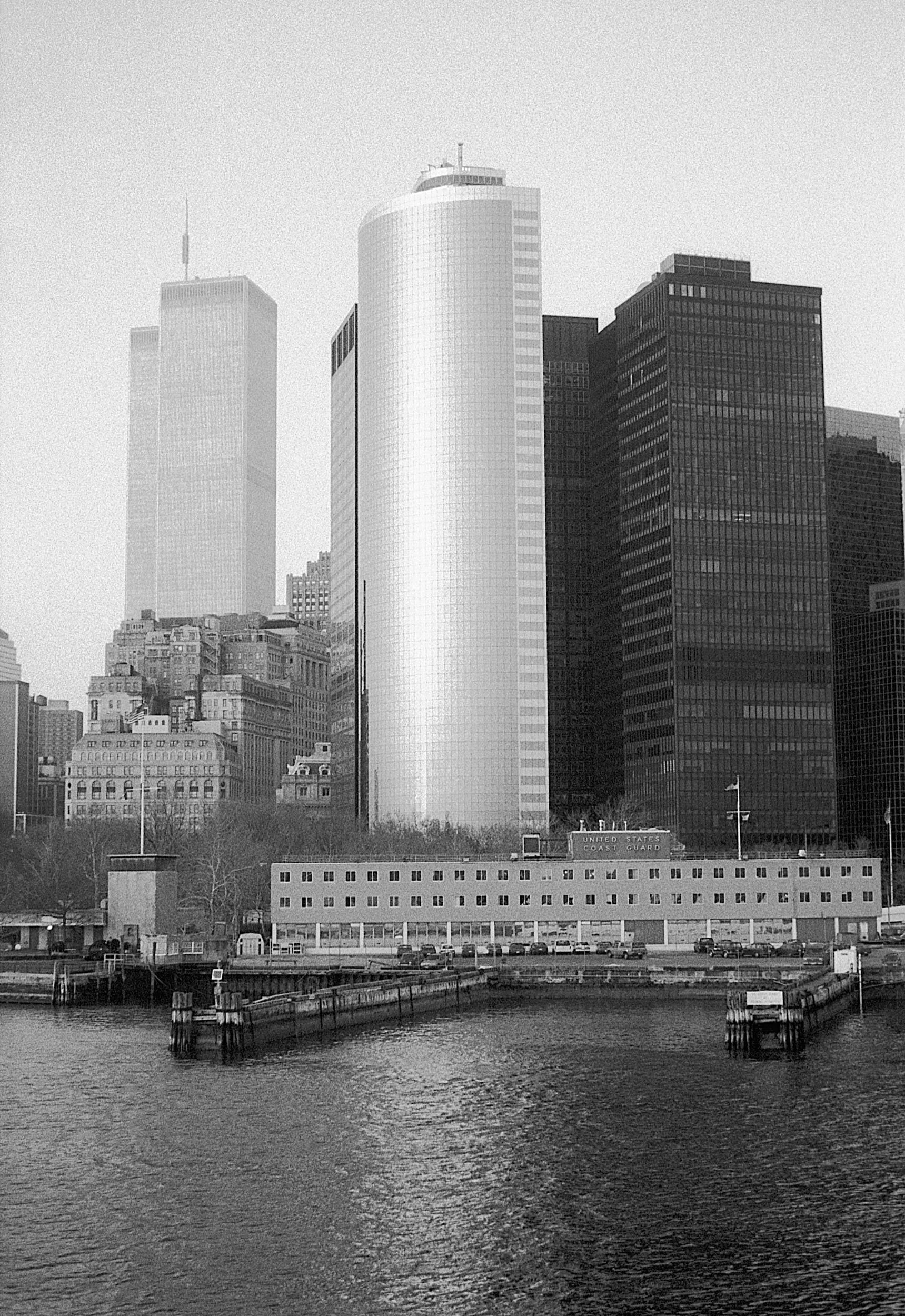 A black and white photograph of a city skyline viewed from across a body of water, featuring tall skyscrapers and buildings, including the United States Coast Guard building.