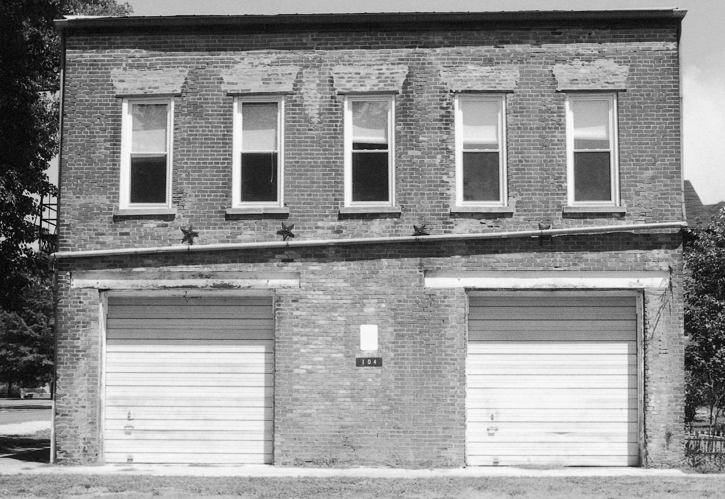 A two-story brick building with four windows on the upper floor and two garage doors on the lower level, with some brickwork in disrepair and a small plaque with the number 104 in the center.