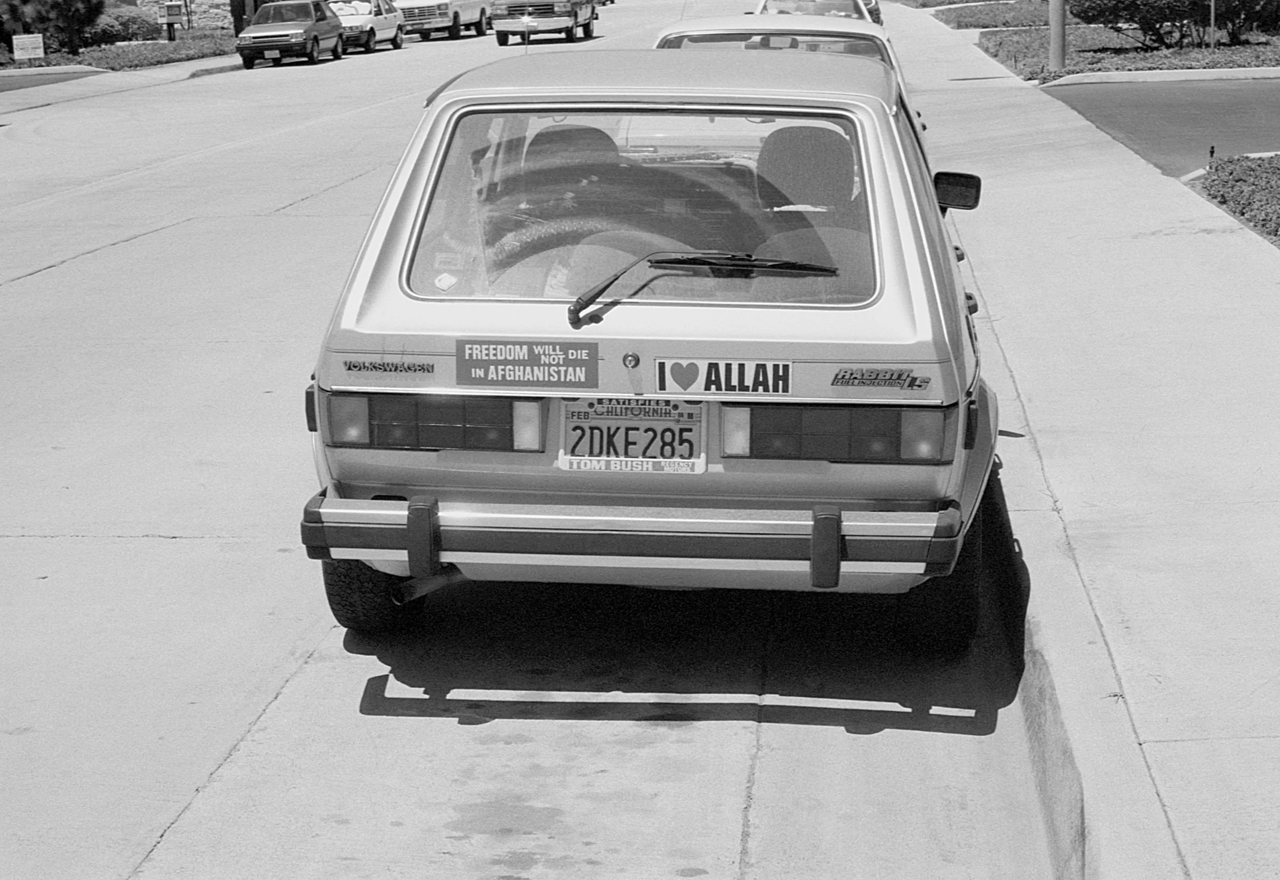 Rear view of a vintage hatchback car with multiple stickers, including one that says 'FREEDOM WILL NOT DIE IN AFGHANISTAN' and another that says 'I ❤️ ALLAH', parked on a sidewalk. Other cars are visible on the street in the background.