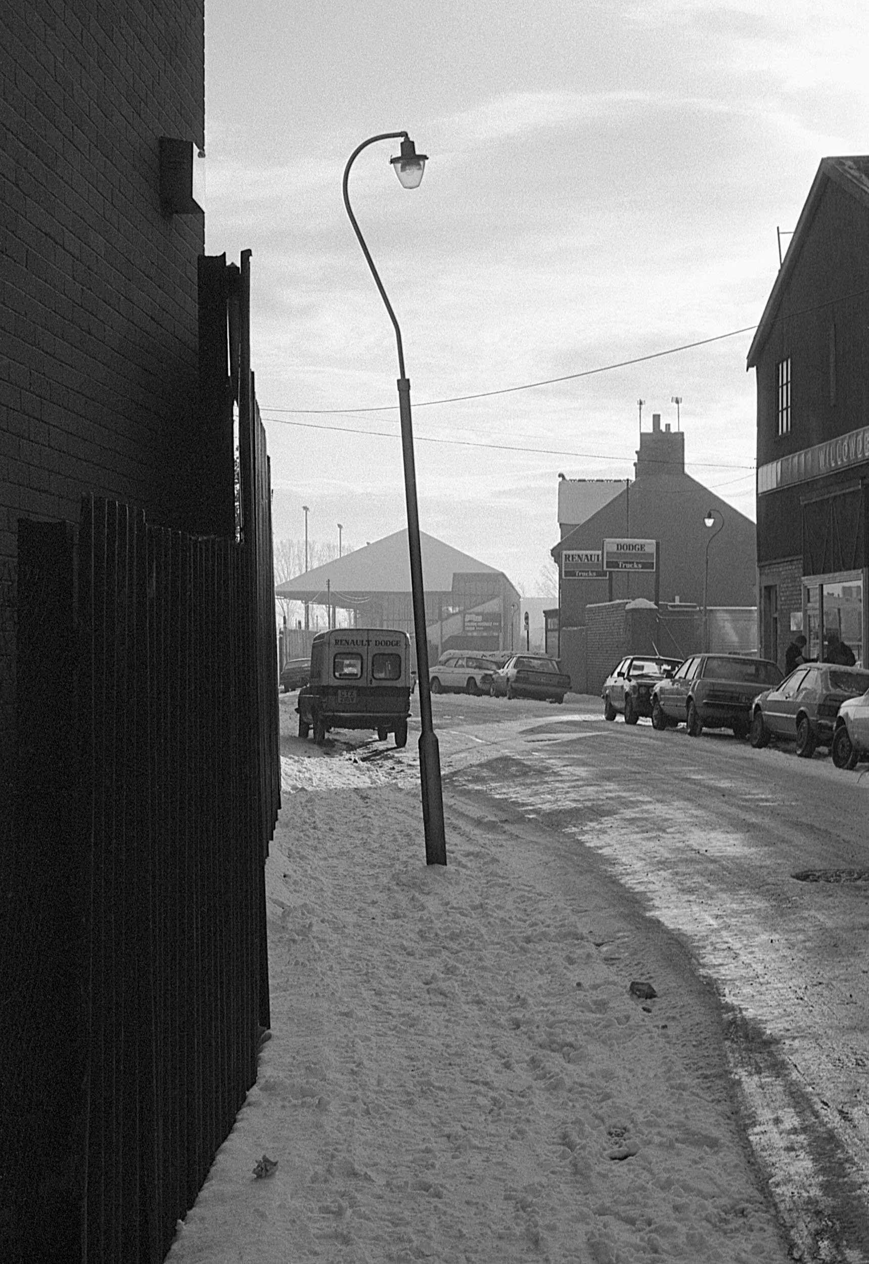 A black and white photograph of a snow-covered street scene with parked cars, a tilted streetlamp, a small Renault Dodge van, buildings, and a sign for Renault and Dodge trucks, under a cloudy sky.