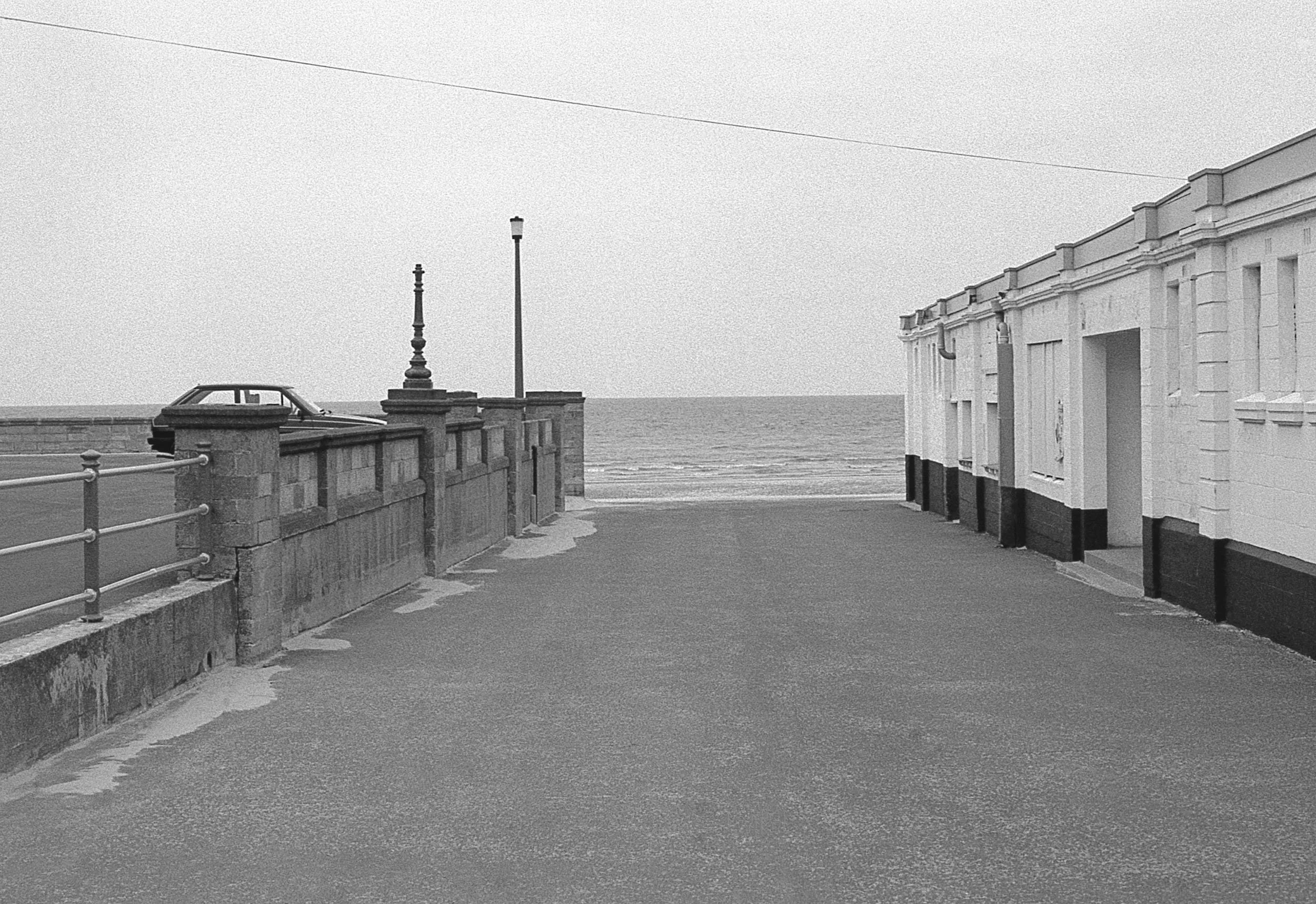 Black and white photo of a seaside promenade with a stone wall and lampposts on the left, and white buildings on the right, leading to the ocean in the background.