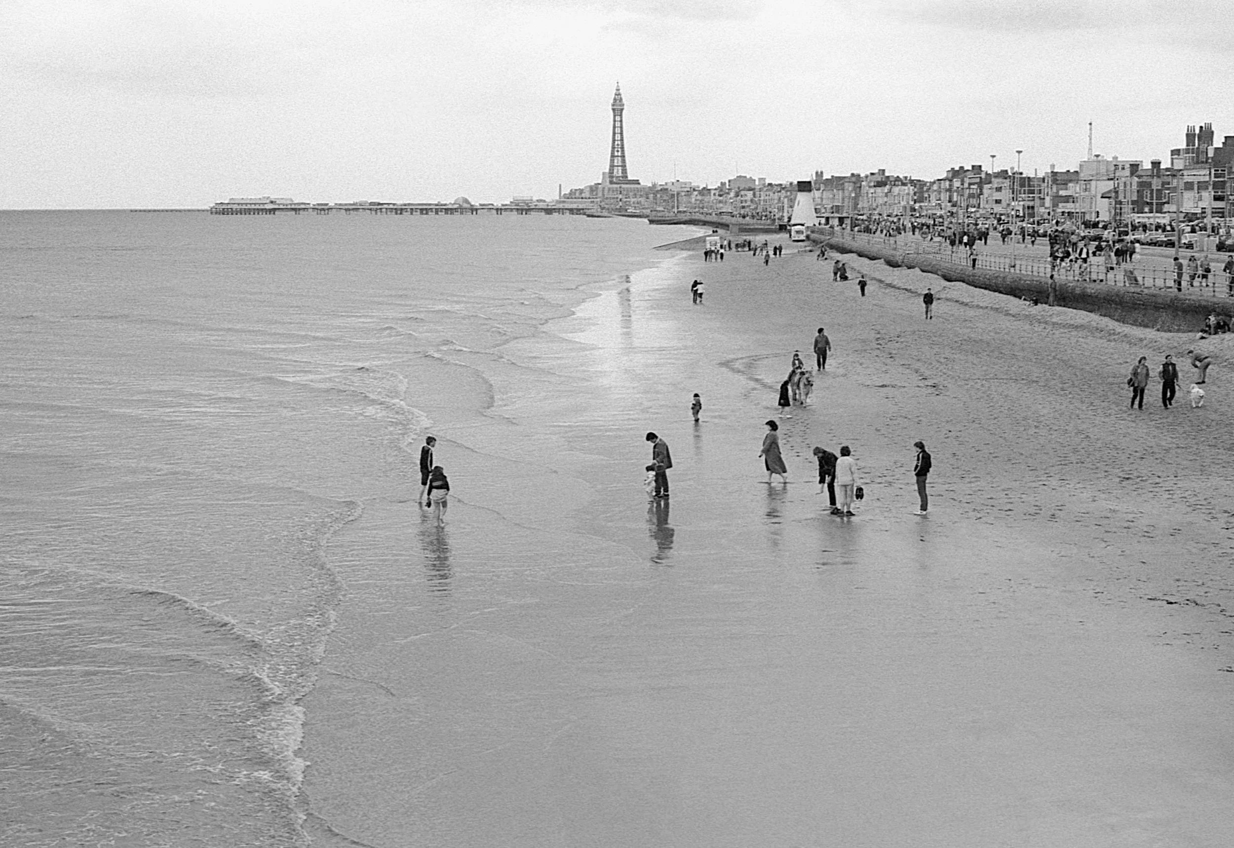 People walking along the shore of a beach with the pier and buildings in the background, photographed in black and white.