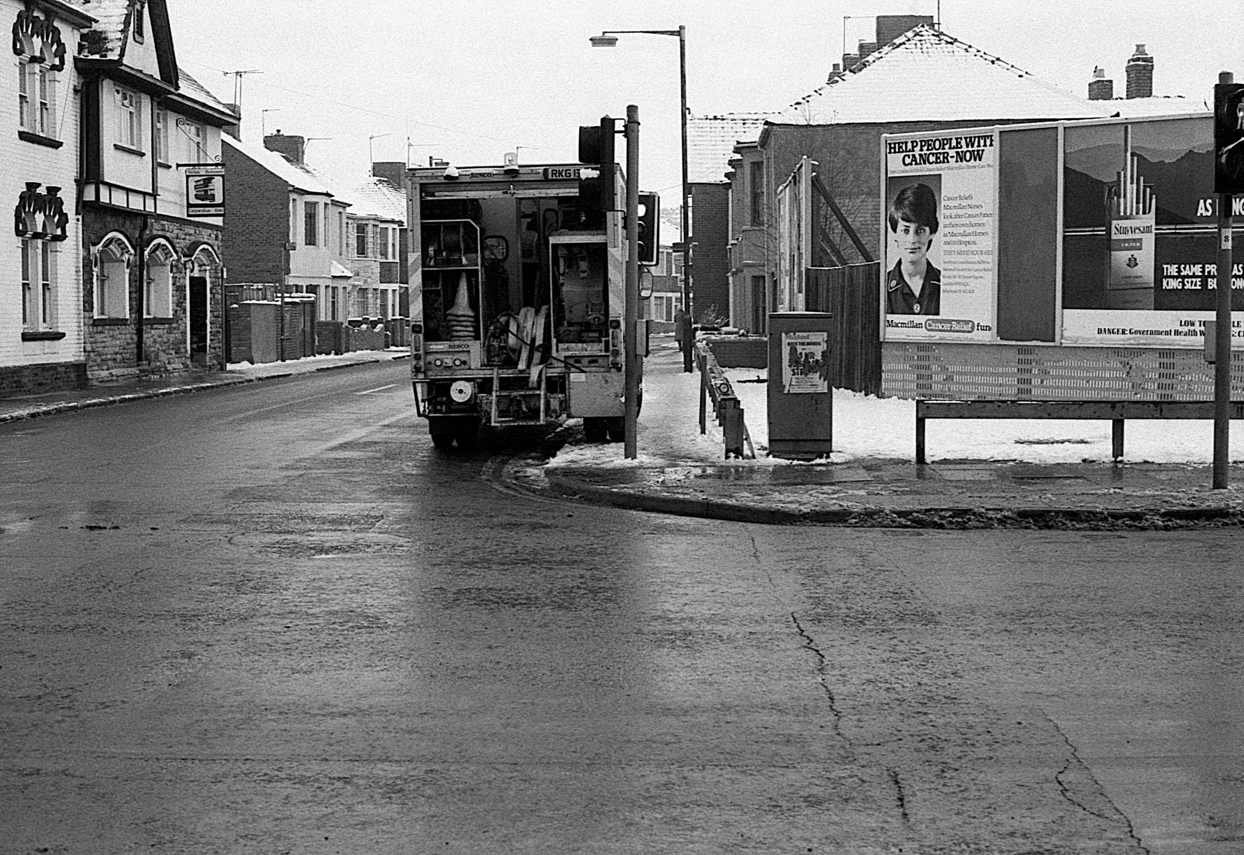 A street corner with a vehicle, possibly a street sweeper, parked near a bus stop. There are houses on the left side and a billboard on the right side with an advertisement for cancer relief. Snow is on the ground and on rooftops, indicating winter w