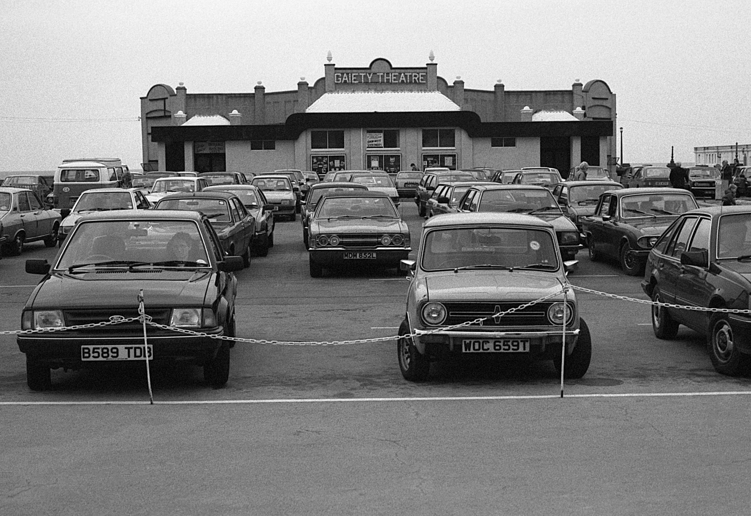Black and white photo of a parking lot in front of a theater with a sign that reads 'Gaiety Theatre'. Several parked cars are visible, with two cars in the foreground separated by a chain barrier.