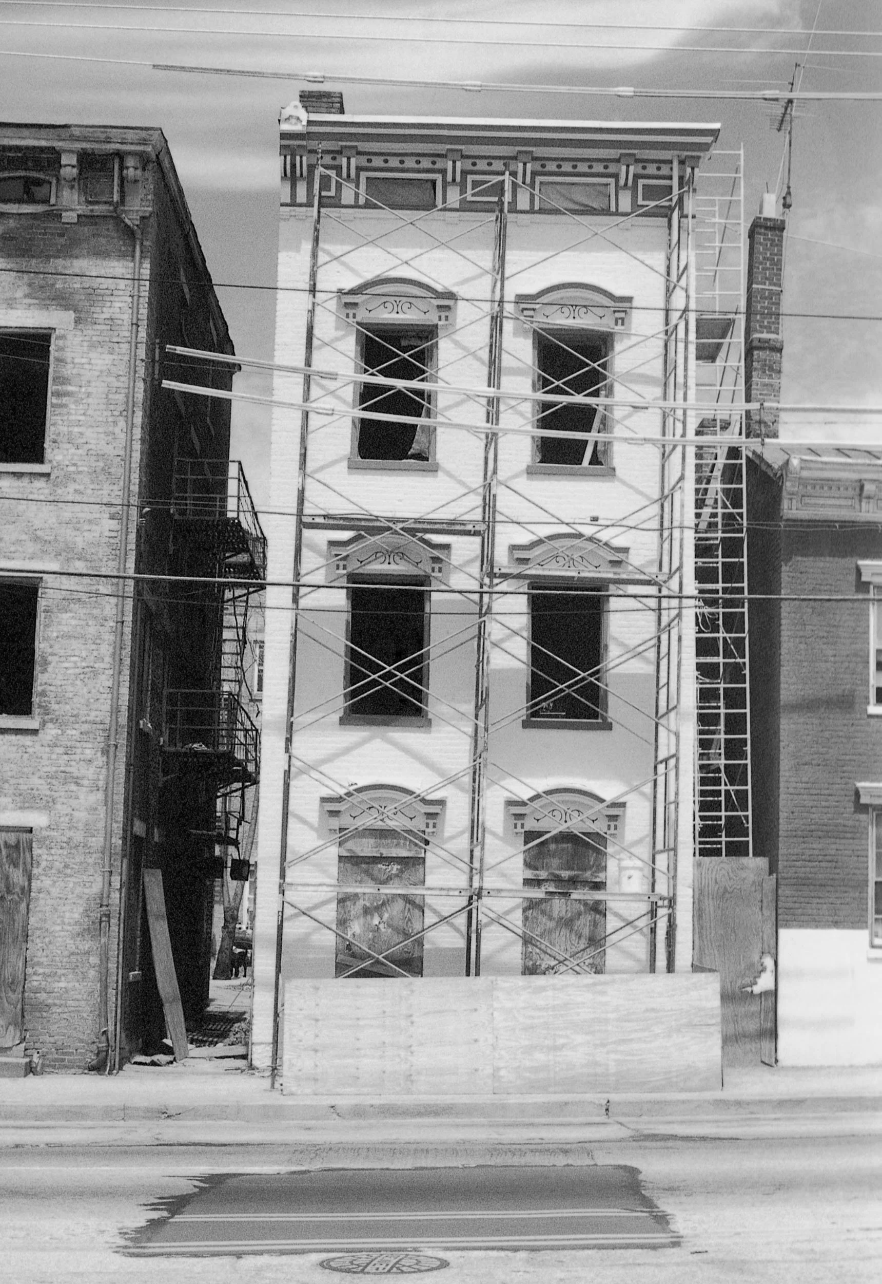 A four-story building under construction or renovation, with scaffolding on the front, and boarded-up ground-floor windows. The building has decorative window trims and an ornate cornice, located between older brick buildings.