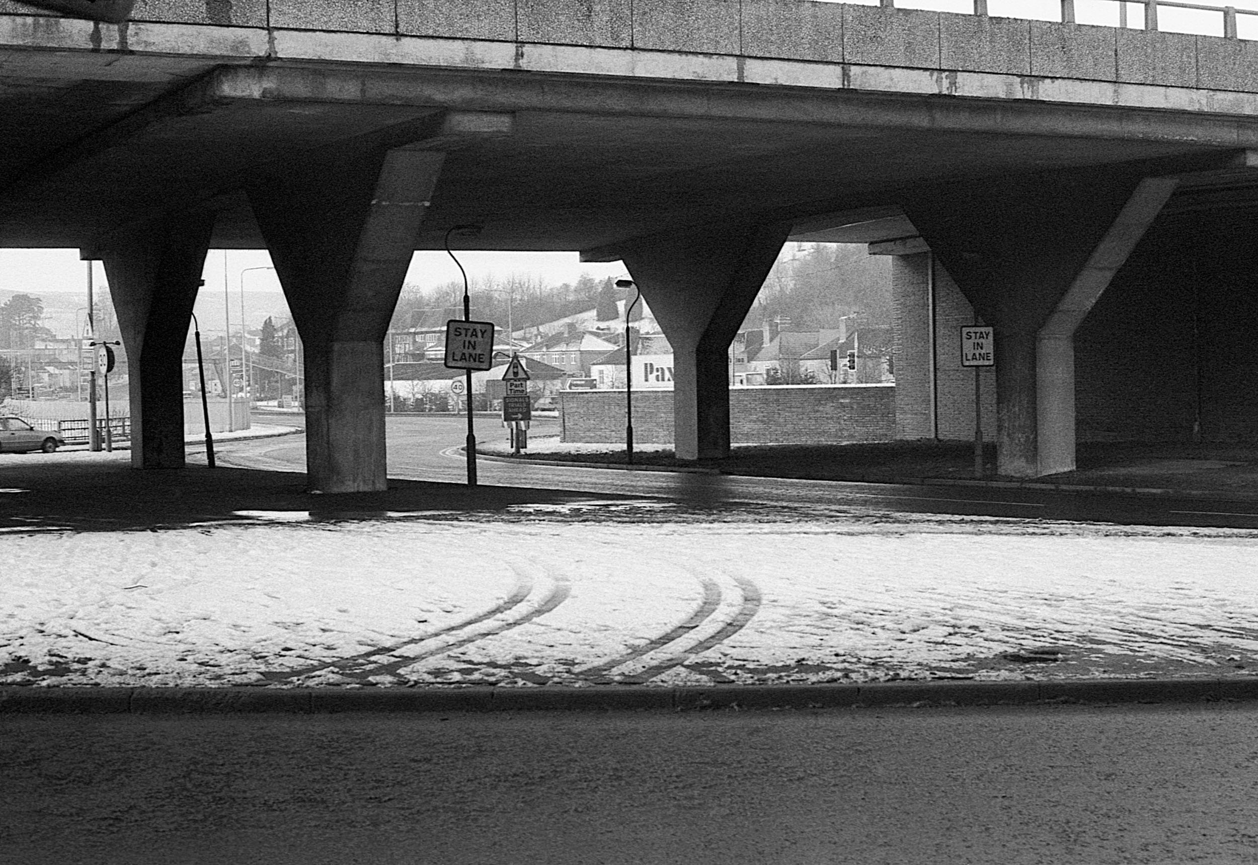 Underpass with snow-covered ground, tire tracks, and multiple signs including 'Stay in Lane' signs, with houses and trees in the background.