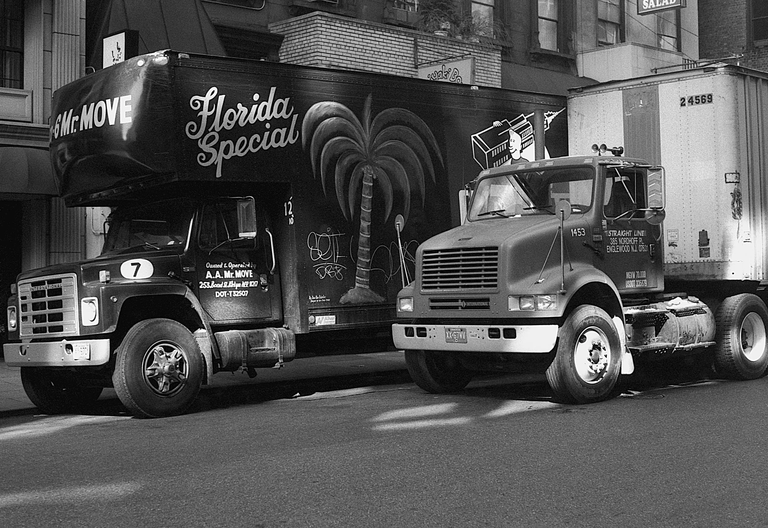 Black and white photograph of two parked trucks on a city street with buildings in the background. The truck on the left has 'Florida Special' written on the side and a palm tree illustration. The right truck is an older model with a boxy front and v