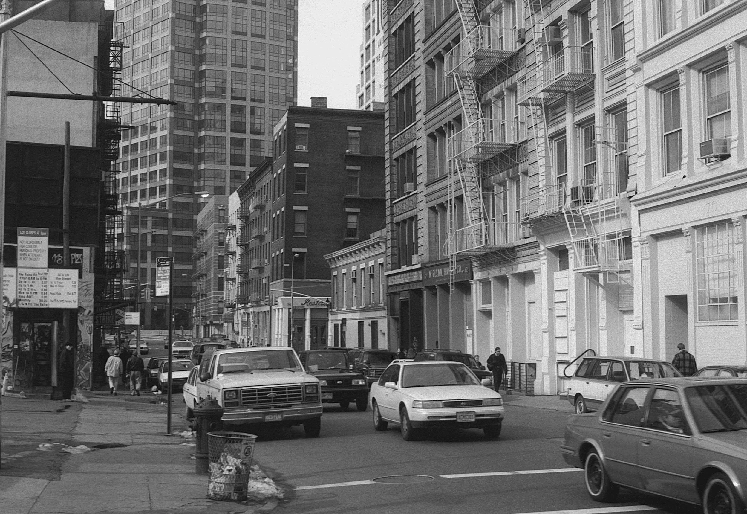 Street scene in an urban area with cars parked along the curb, pedestrians walking, and tall buildings, some with fire escapes, lining the street.