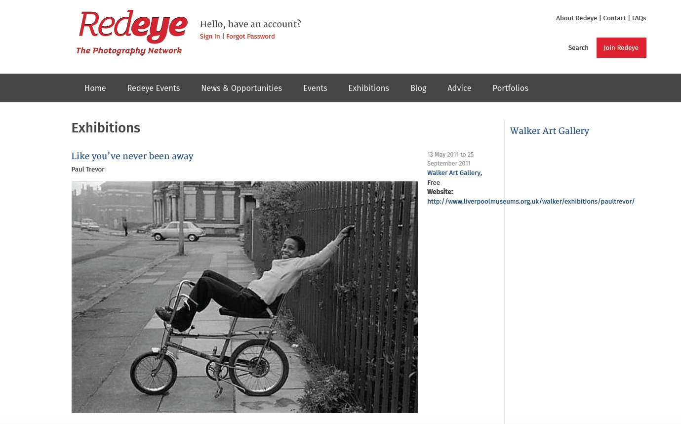 Black and white photo of a person riding a bicycle on the sidewalk, smiling and reaching out to touch a fence on a city street.