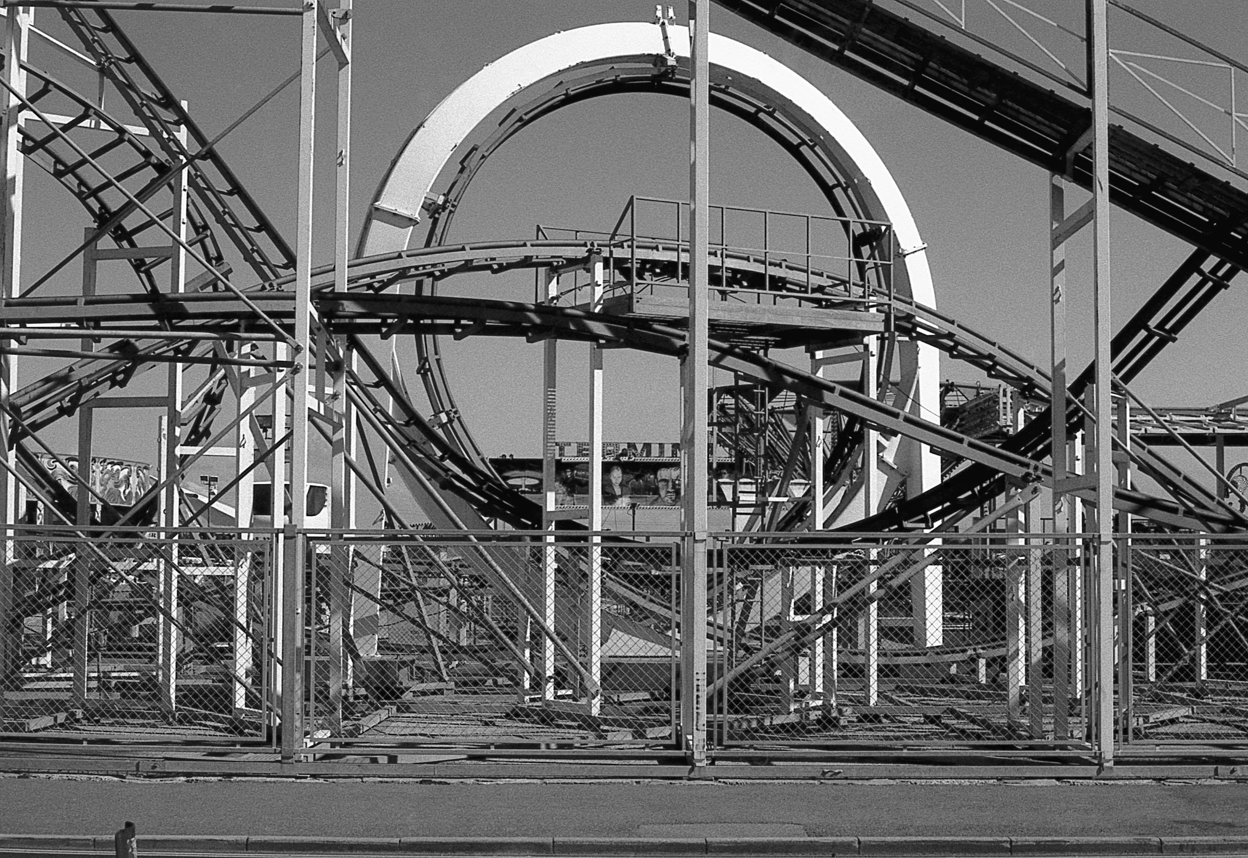 Black and white photo of a roller coaster at an amusement park with metal track tunnels and fencing in the foreground.