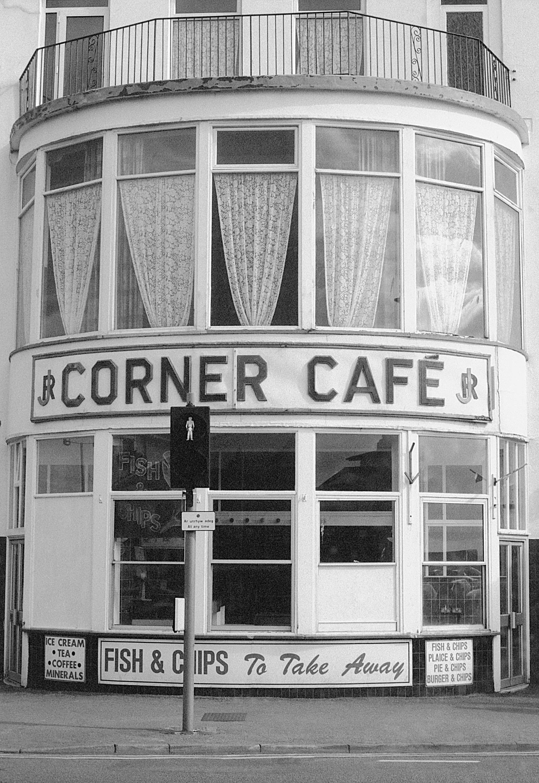 Black and white photo of a corner café with large curved glass windows, a sign that reads 'Corner Café', and a smaller sign advertising fish and chips to go. There is a pedestrian crossing signal in front and a sidewalk with street markings.