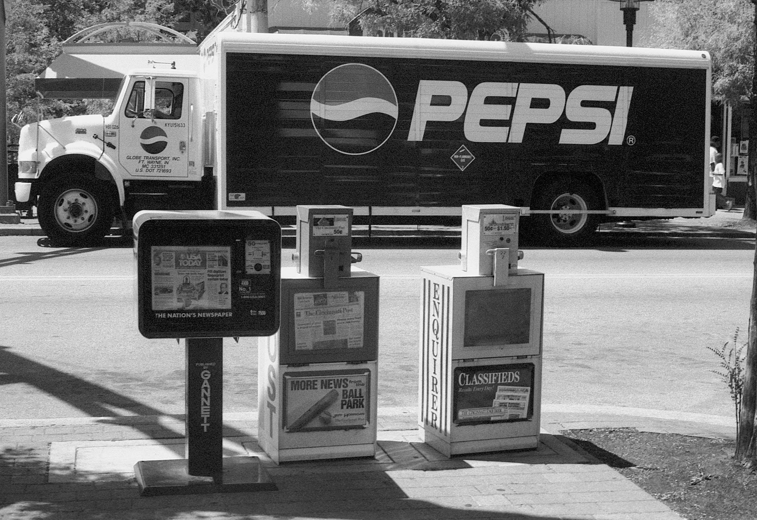 A newspaper vending machine and a magazine rack outside on the sidewalk in front of a street with a Pepsi delivery truck passing by.