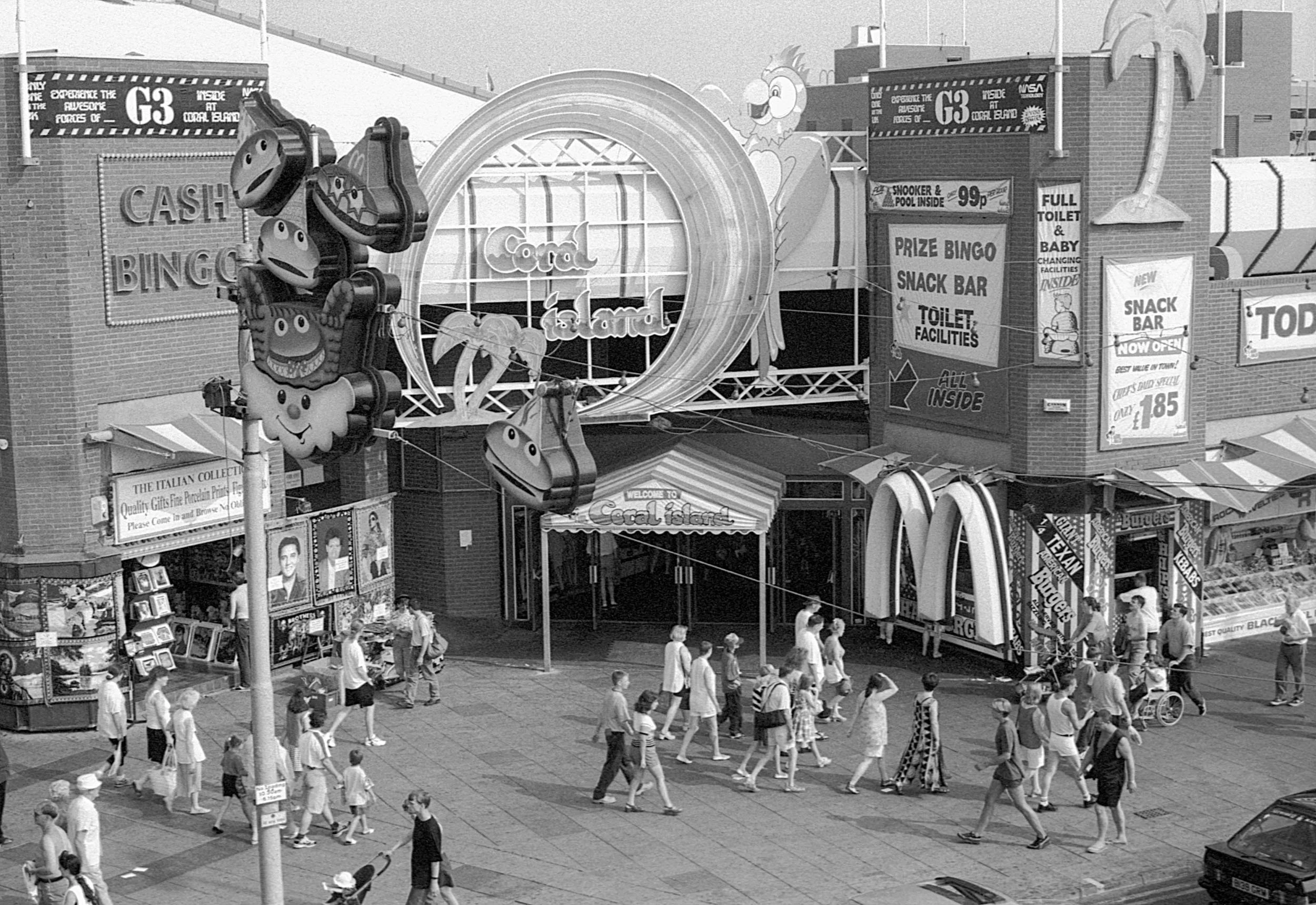 People walking outside a shopping mall with amusement park rides and signs for food and entertainment, including a large sign for Coral Island and murals of big animals.