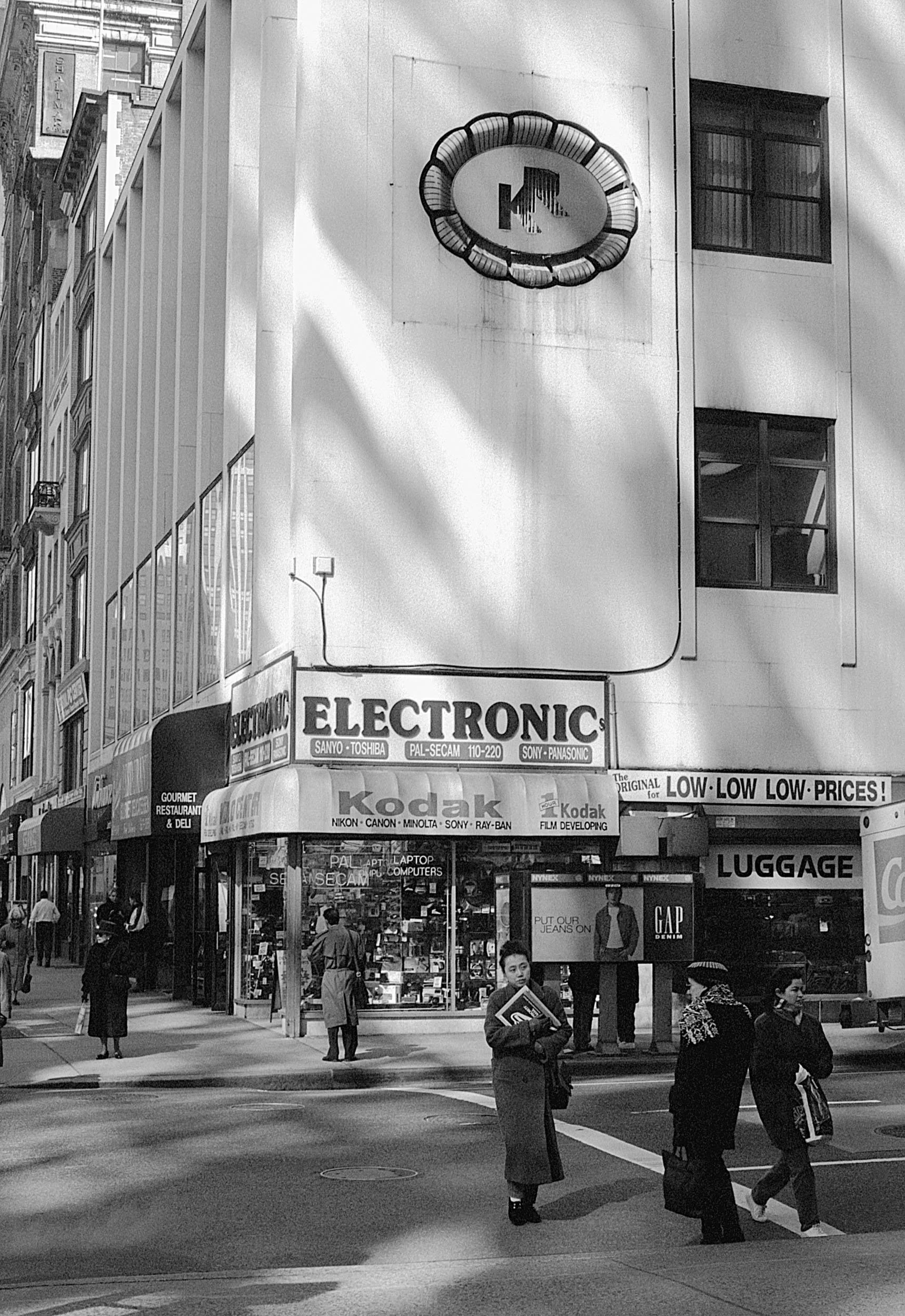 Street scene in an urban area with pedestrians crossing the street, storefronts with signs for electronics and camera stores, and tall buildings in the background.