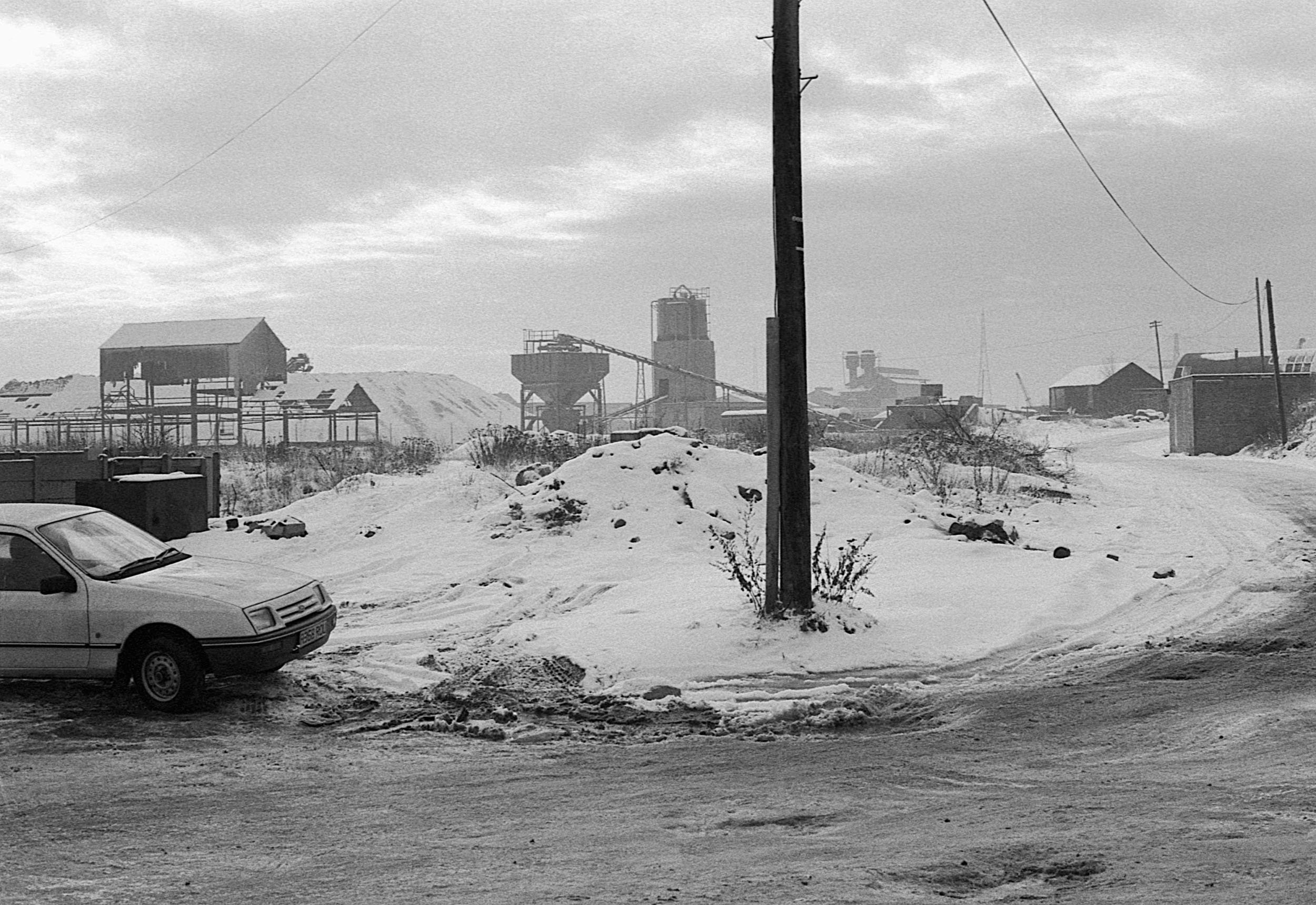 Snow-covered industrial landscape with a car parked in the foreground and industrial buildings with silos and structures in the background.