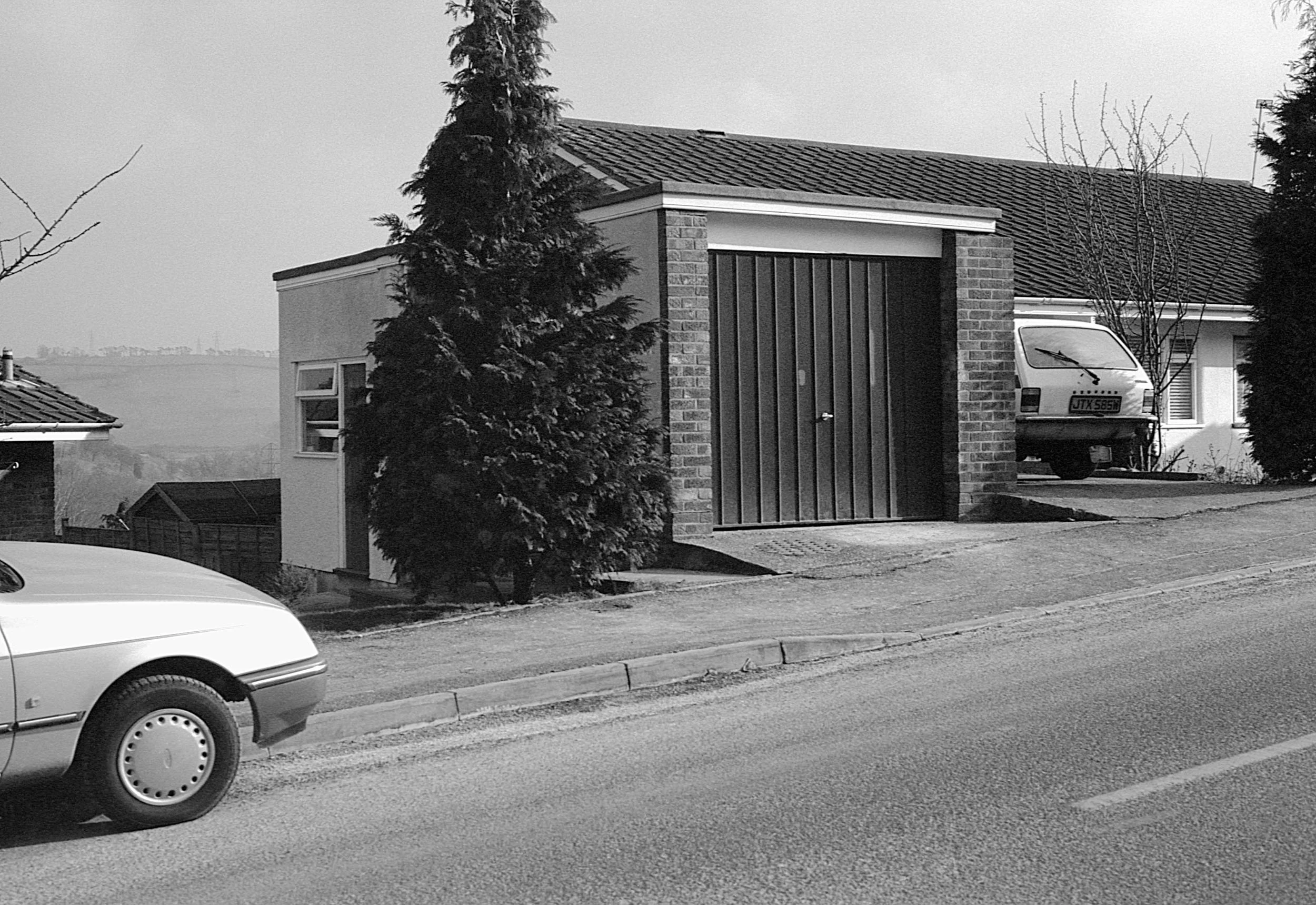 A street scene in black and white showing a portion of a car parked on the left, a house with a garage in the center, and another car parked behind the garage. There are trees and a distant landscape in the background.
