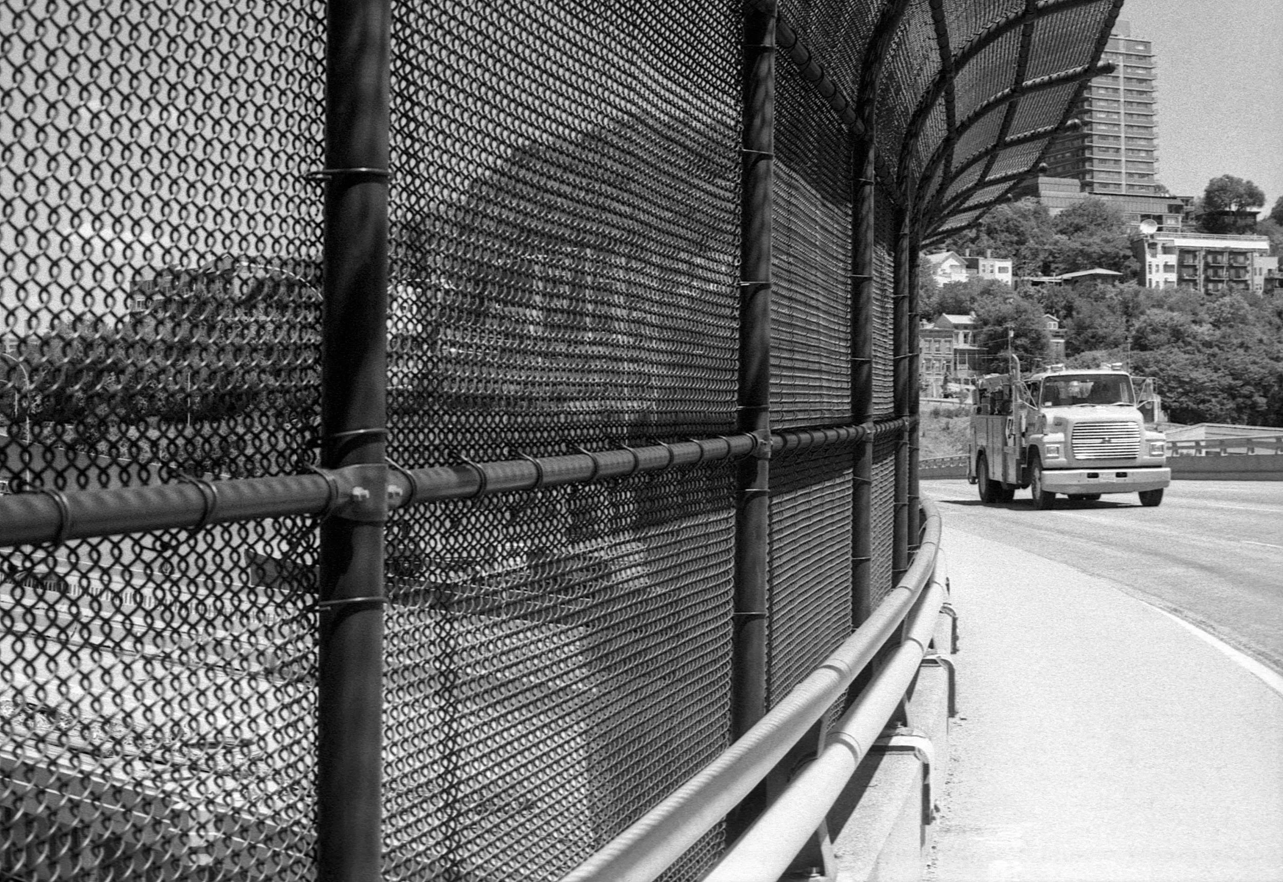 A highway with a chain-link fence on the left side and a truck driving on the right side, with buildings and trees in the background.