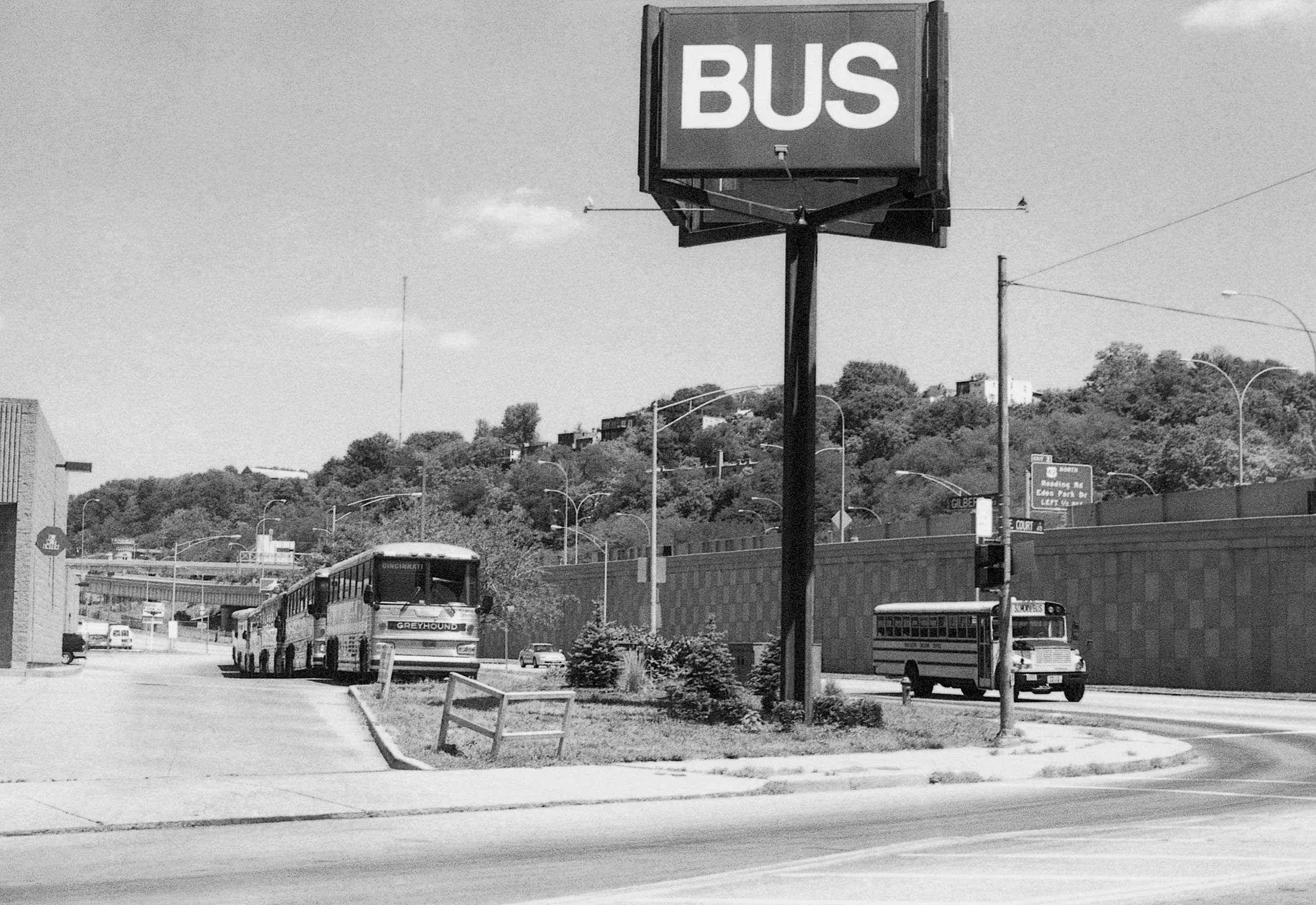 Black and white photo of a bus stop area with a large sign that says 'BUS' on a tall pole. There are buses and cars on the road, with a hillside filled with trees in the background. A school bus labeled 'SCHOOL BUS' is visible on the right side.