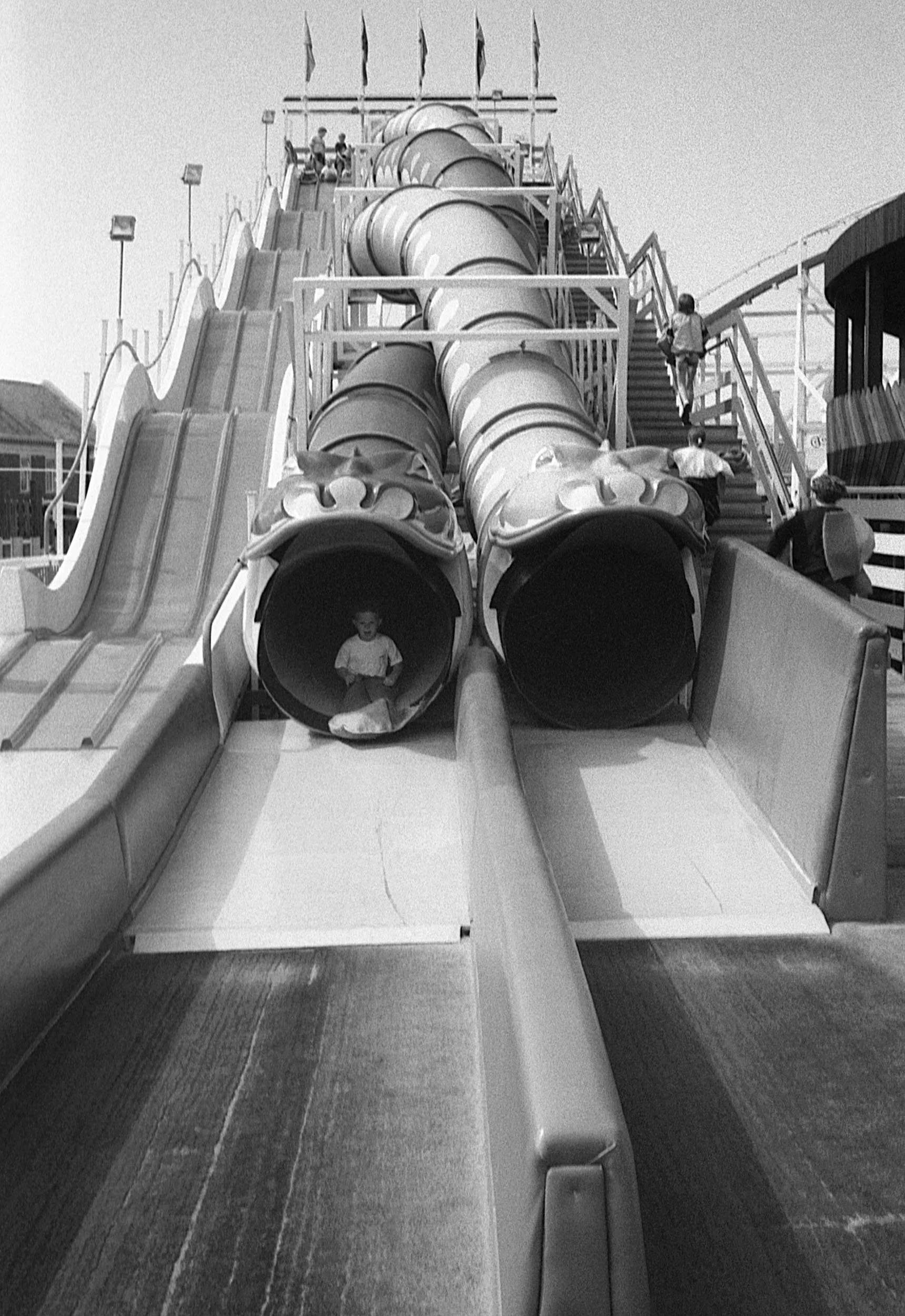 A black and white photo of an amusement park slide with multiple twisting tubes. Children are sliding down and others are walking up the stairs beside the slide.