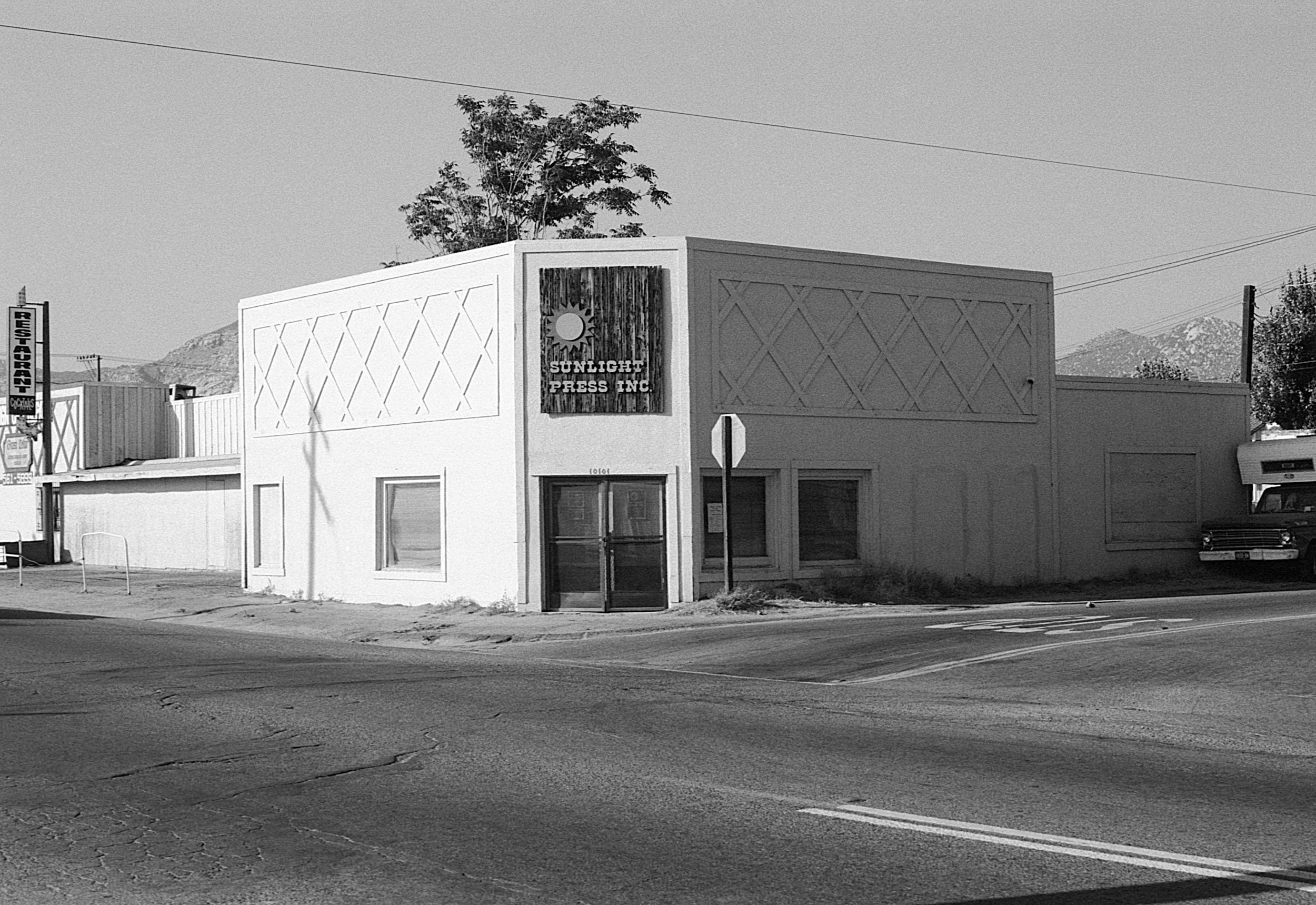 A black and white photo of a corner building with a sign that reads 'Sunlight Press Inc.' next to a stop sign. The building has a decorative upper wall and three windows on the lower level, with one door. The street in front has visible cracks, and t