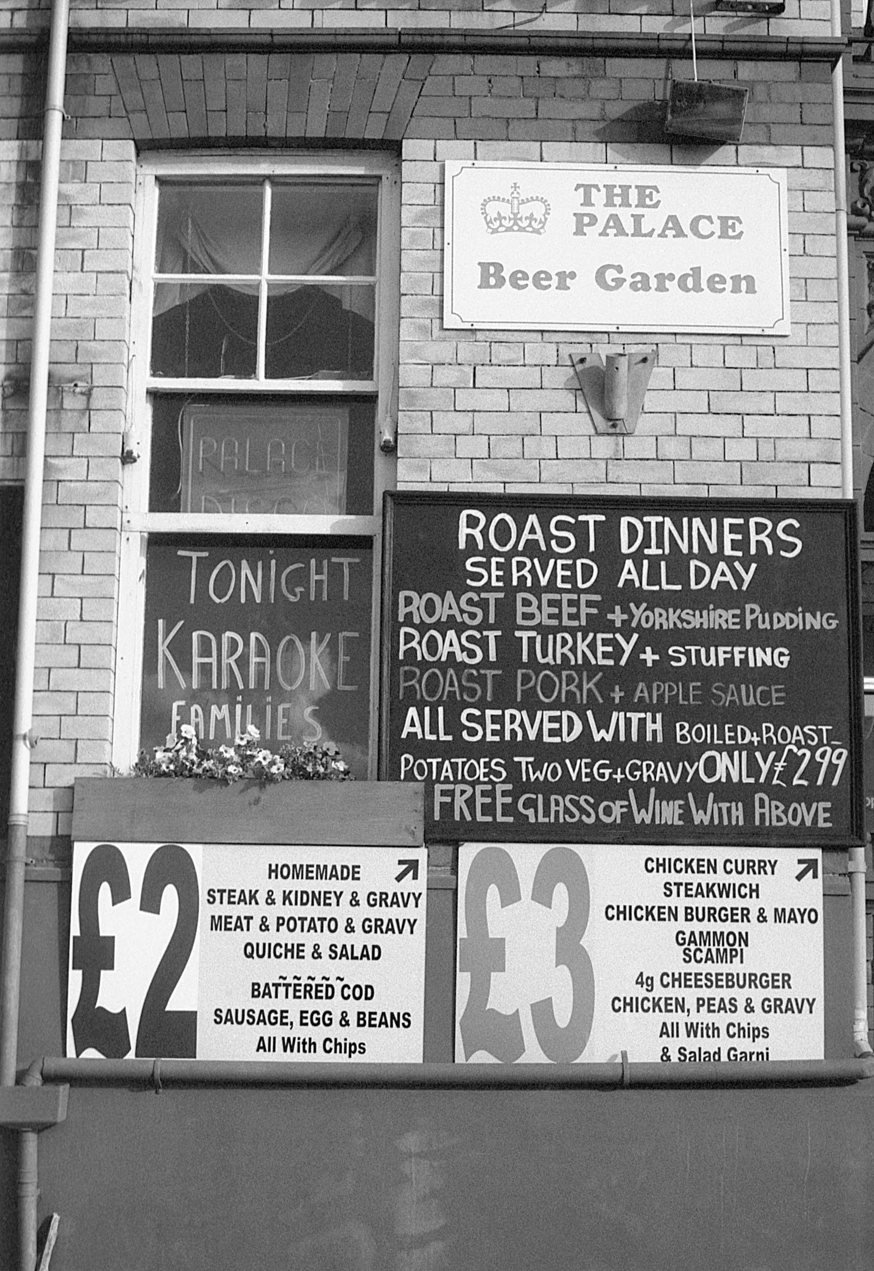 Sign for The Palace Beer Garden advertising roast dinners served all day, including roast beef, turkey, and pork, with potatoes, vegetables, and gravy, plus a free glass of wine. Menu prices are £2 for homemade dishes and £3 for chicken dishes, with 