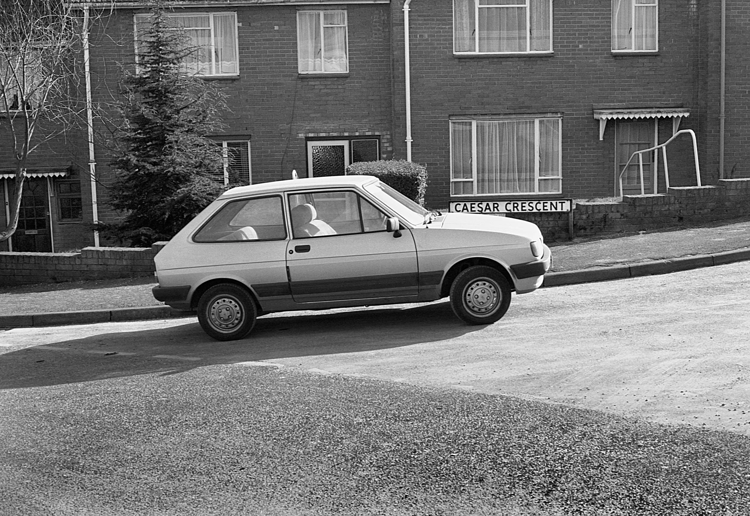 A vintage compact car parked on the side of the street in front of an apartment building. The building has brick walls, several windows, and a sign that reads "Caesar Crescent."